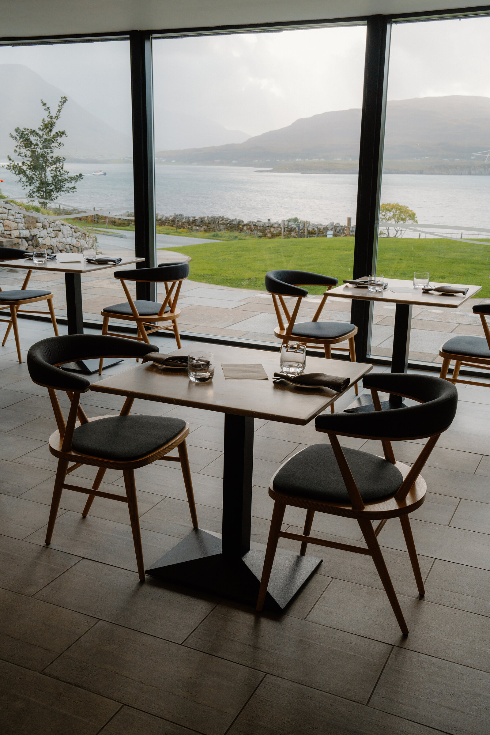 A table set for two overlooks a loch and mountains through floor-to-ceiling windows at the Isle of Raasay Distillery.