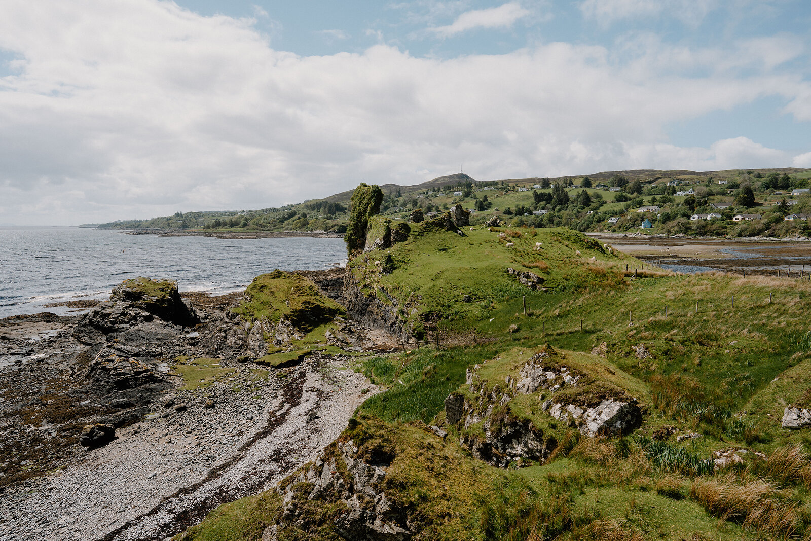 A ruined castle's broken walls stand on a grassy headland above the shore, a village visible along the coast on the Isle of Skye.