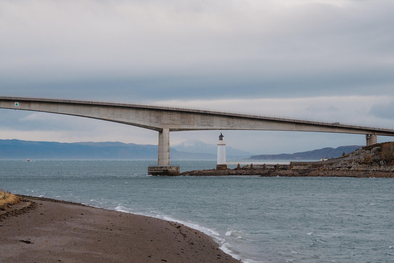 The Skye Bridge sweeps across the water past a small lighthouse, a shingle beach in the foreground.