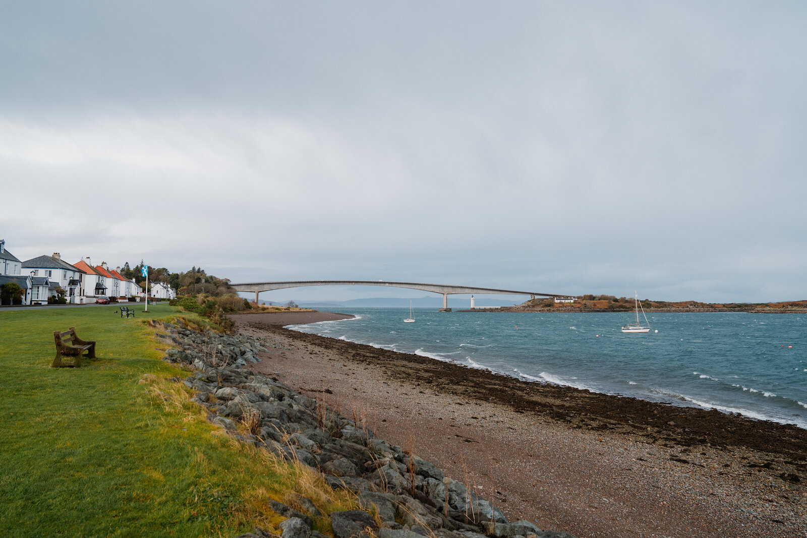 A bench sits on a grassy shore looking out to the Skye Bridge and a moored sailboat.