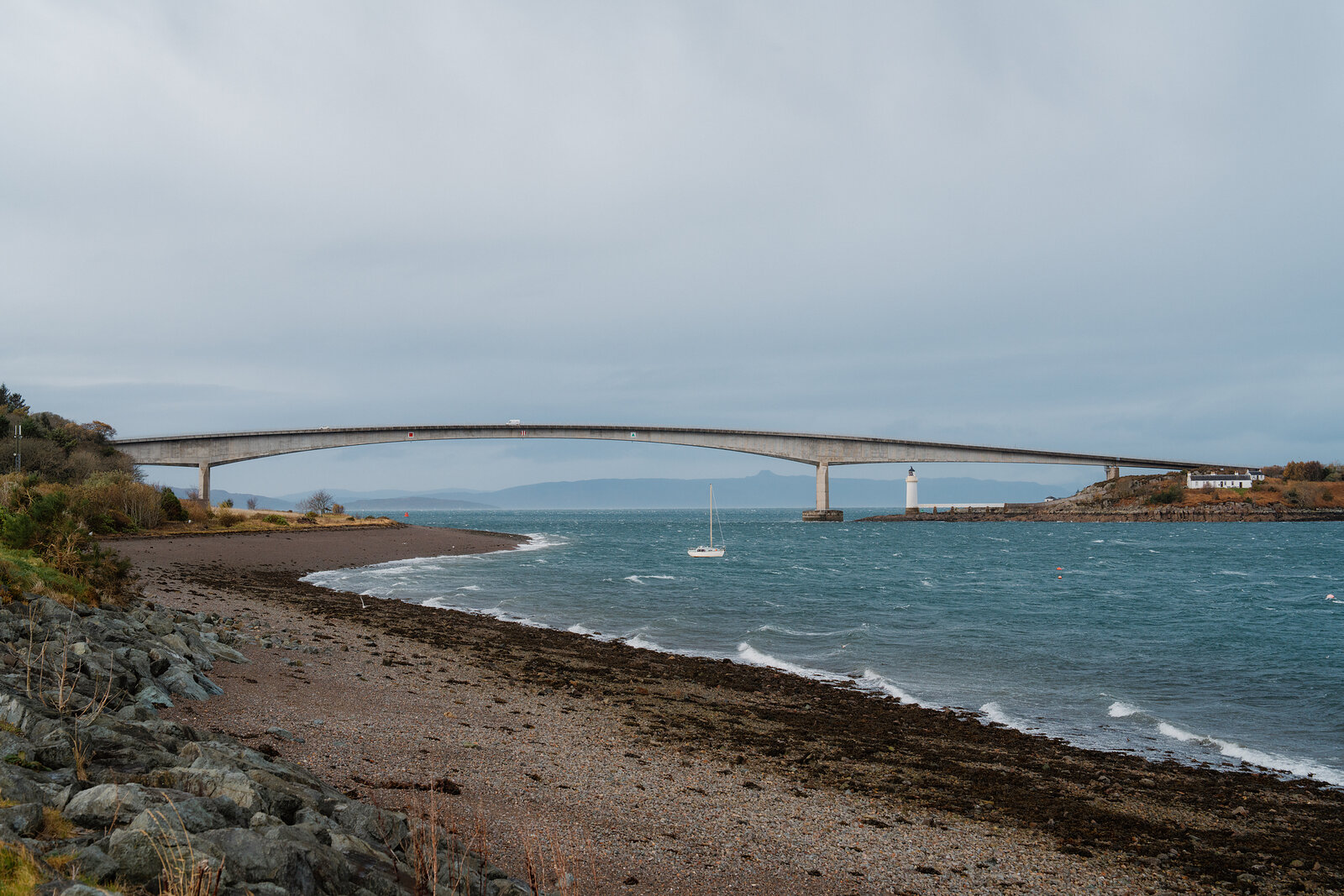 The Skye Bridge stretches across the water past a lighthouse, a sailboat anchored below on the Isle of Skye.