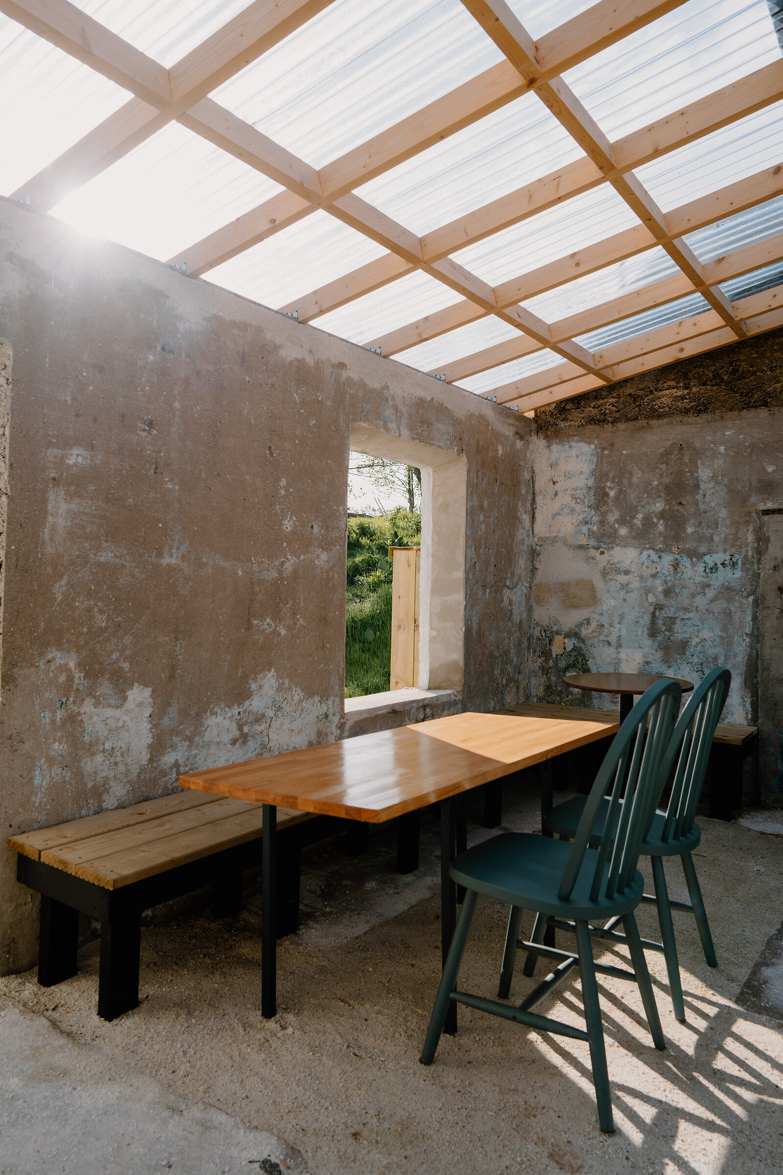 A wooden table and green chairs sit beneath a glass roof in a ruined stone building at Lean to Coffee.