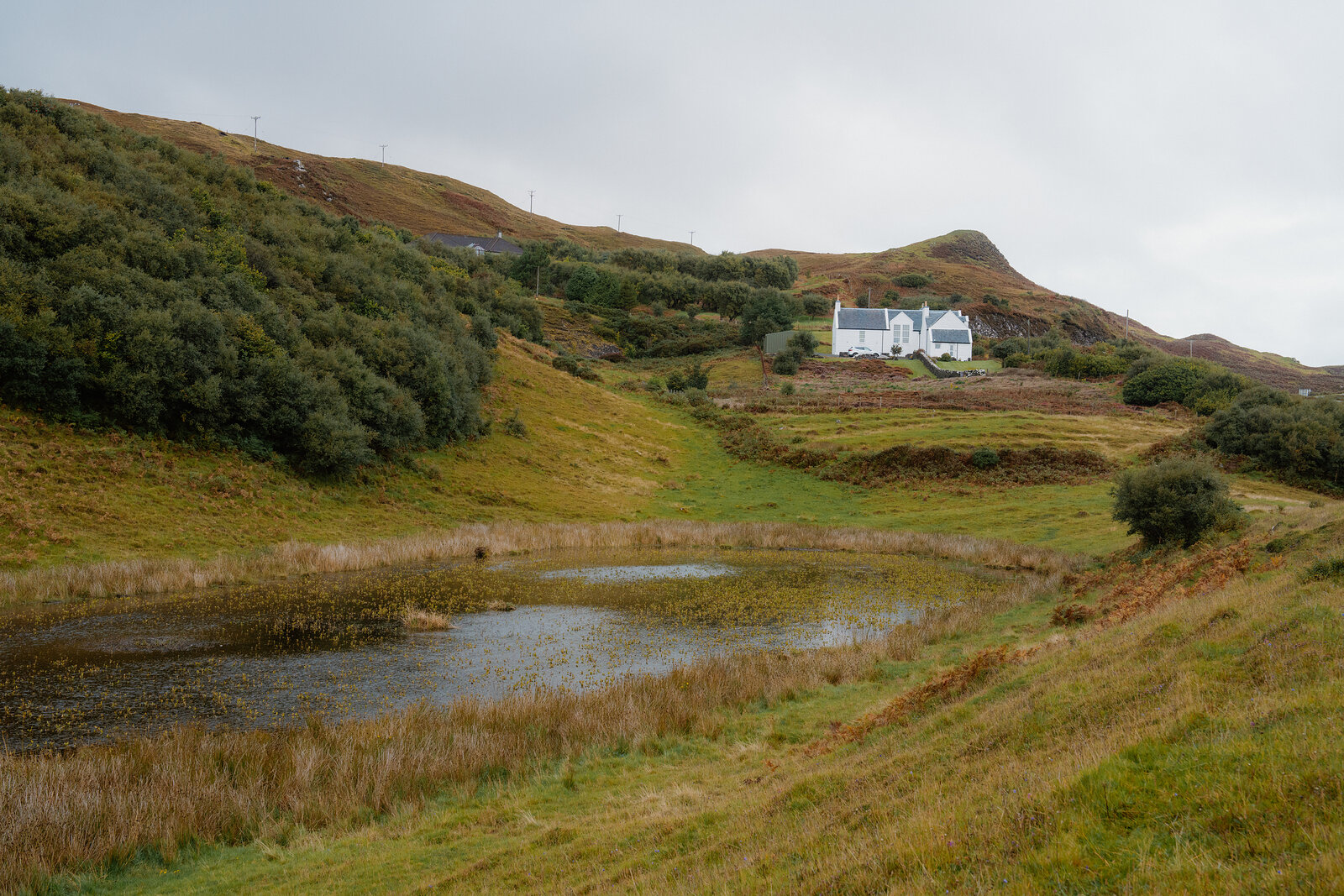 A white house sits on a grassy hillside above a still pond on the Isle of Skye.