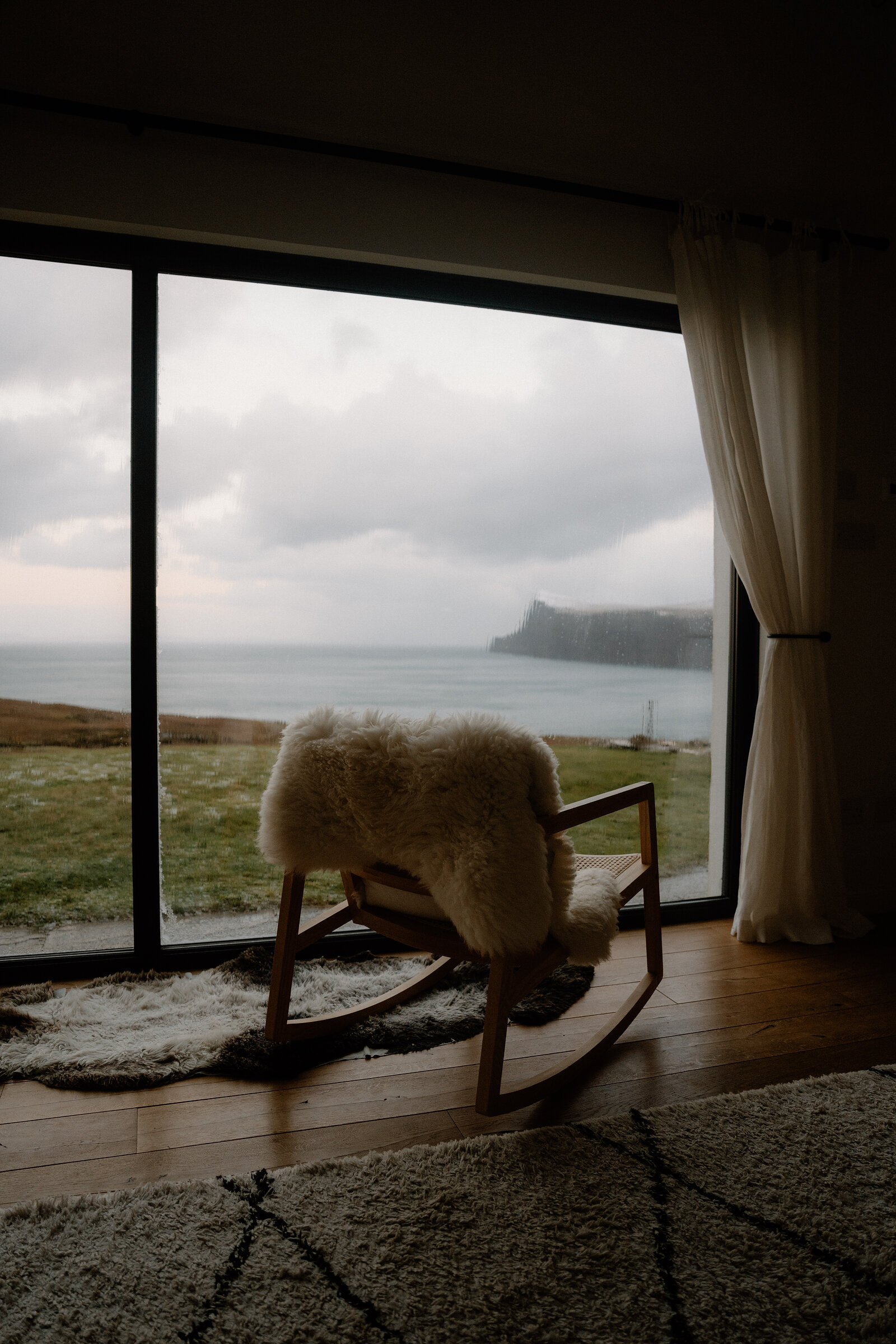 A sheepskin-draped rocking chair faces a window overlooking a cliff and sea on the Isle of Skye.