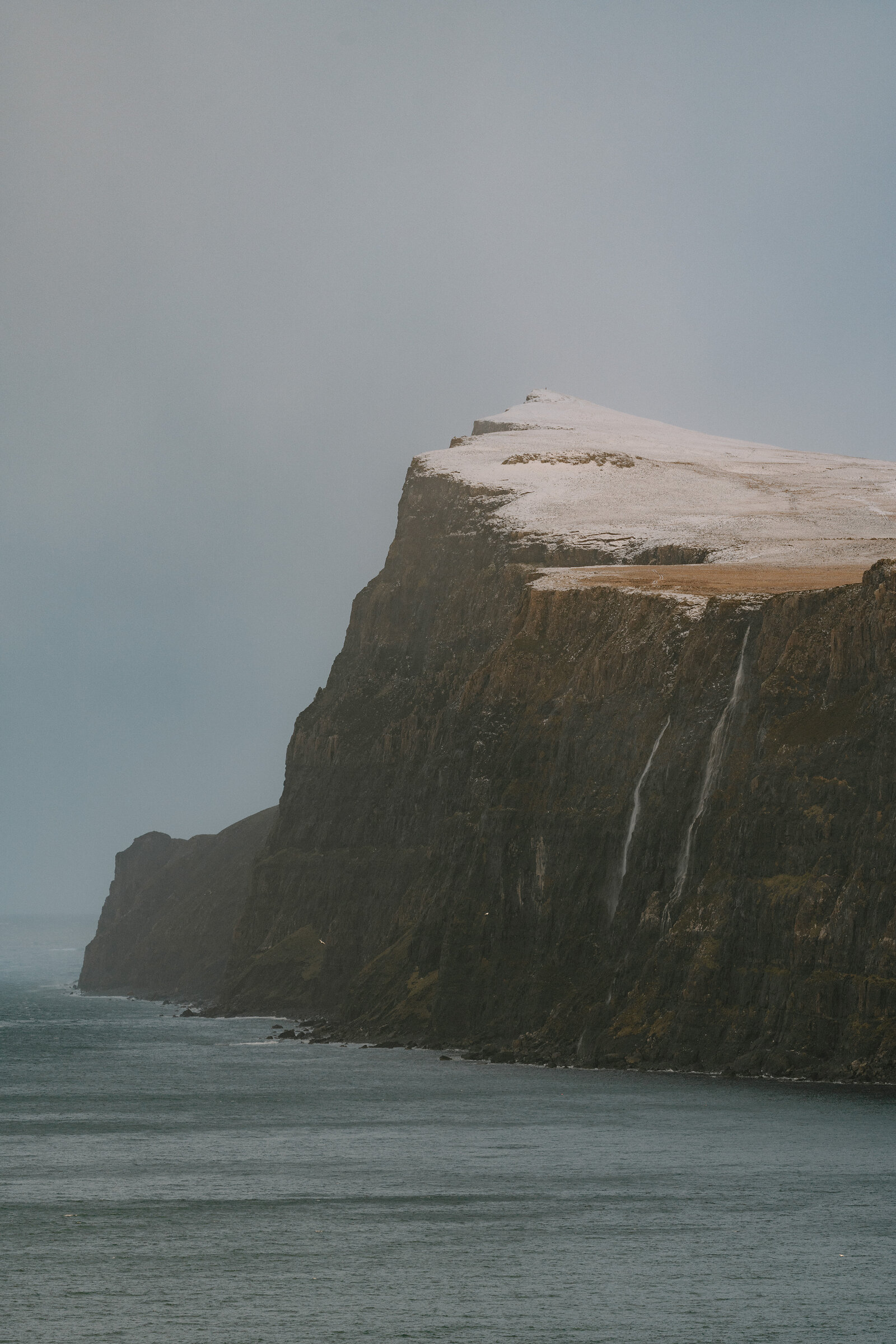 Snow caps a sea cliff on the Isle of Skye, waterfalls streaking down its dark rock face.