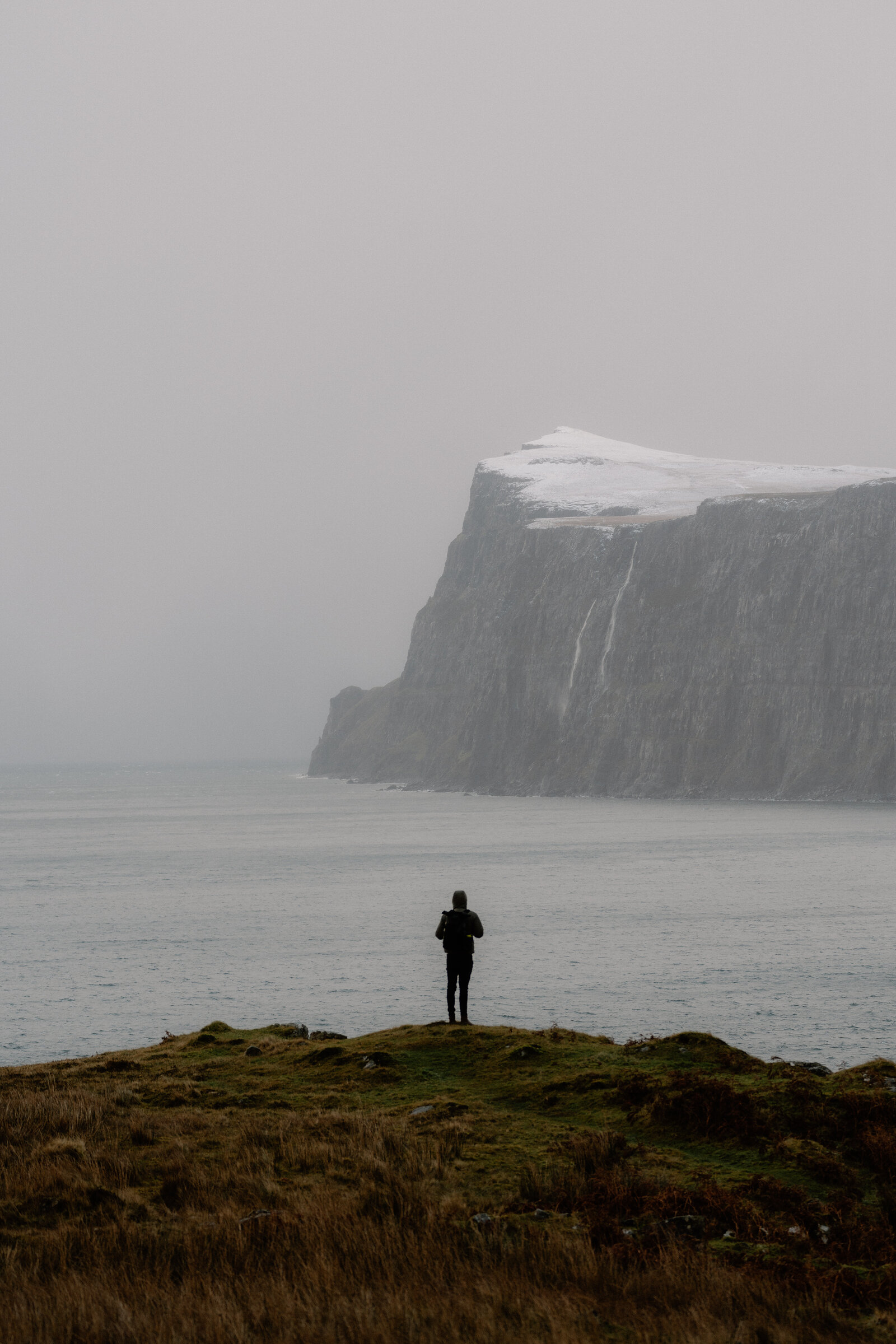 A figure stands on a headland watching a snow-dusted sea cliff and waterfall on the Isle of Skye.