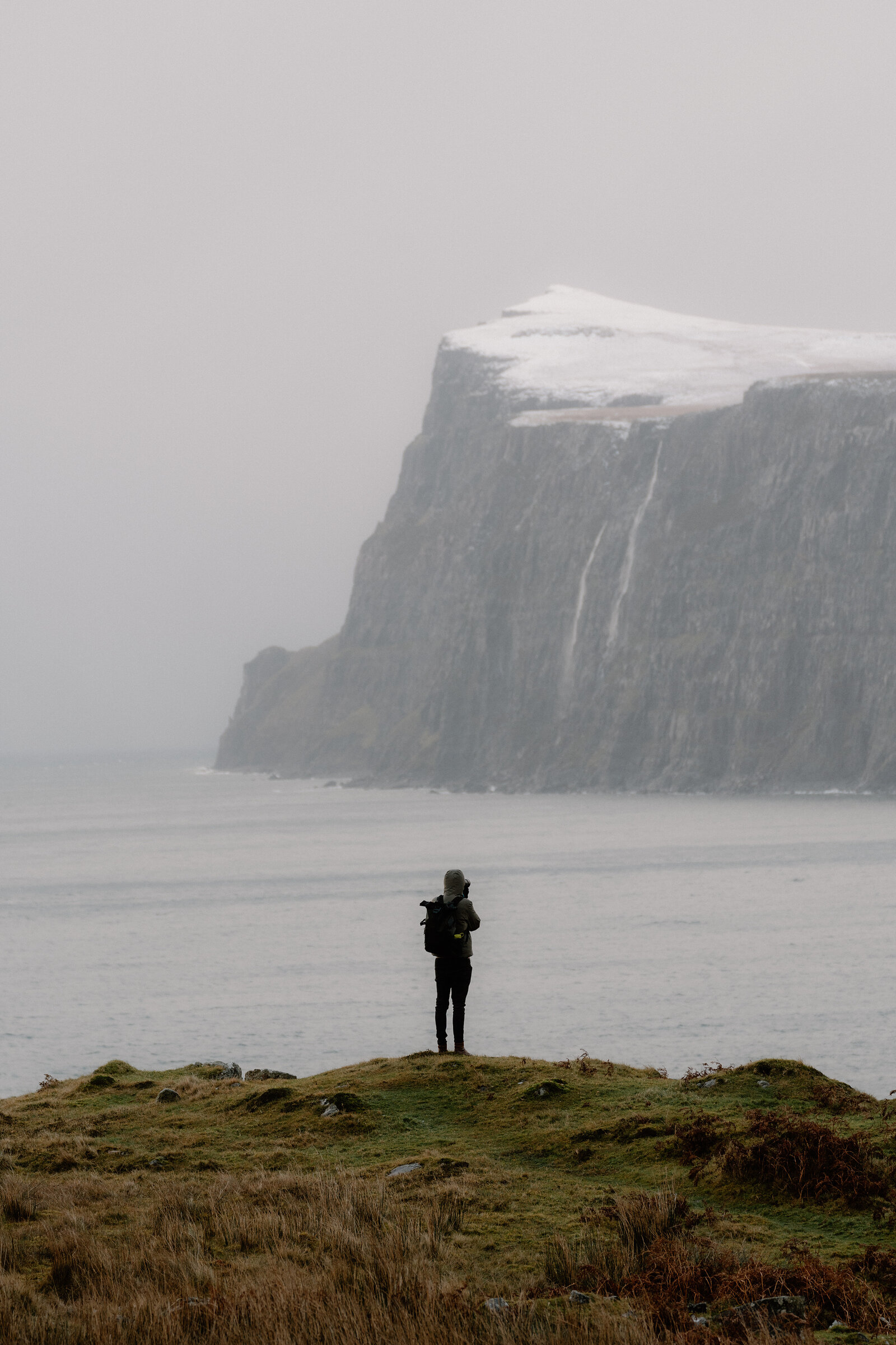 A figure in a hooded jacket stands on a grassy headland facing a snow-capped cliff on the Isle of Skye.