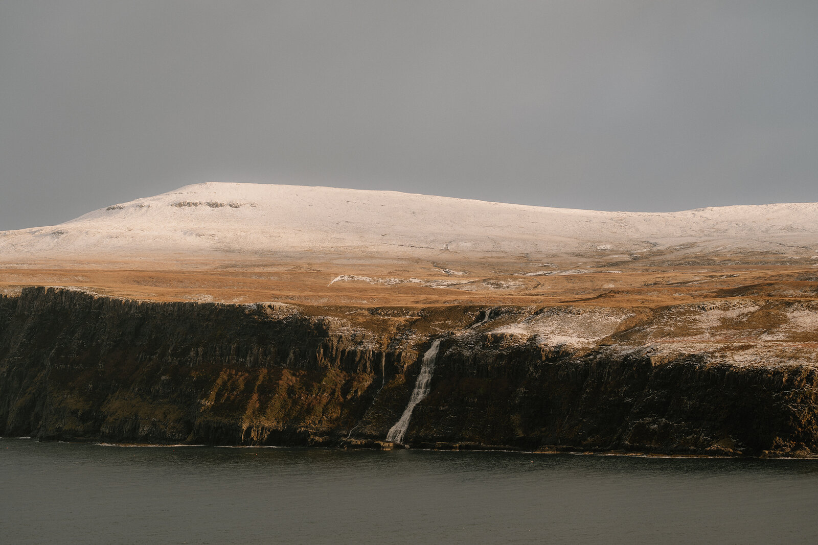 A waterfall streaks down a dark sea cliff below snow-dusted moorland on the Isle of Skye.