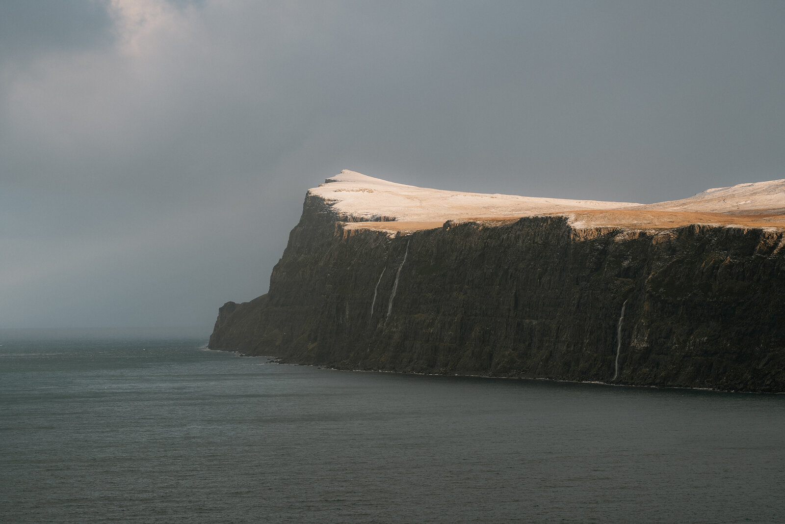 Waterfalls streak down a dark sea cliff capped with snow on the Isle of Skye.
