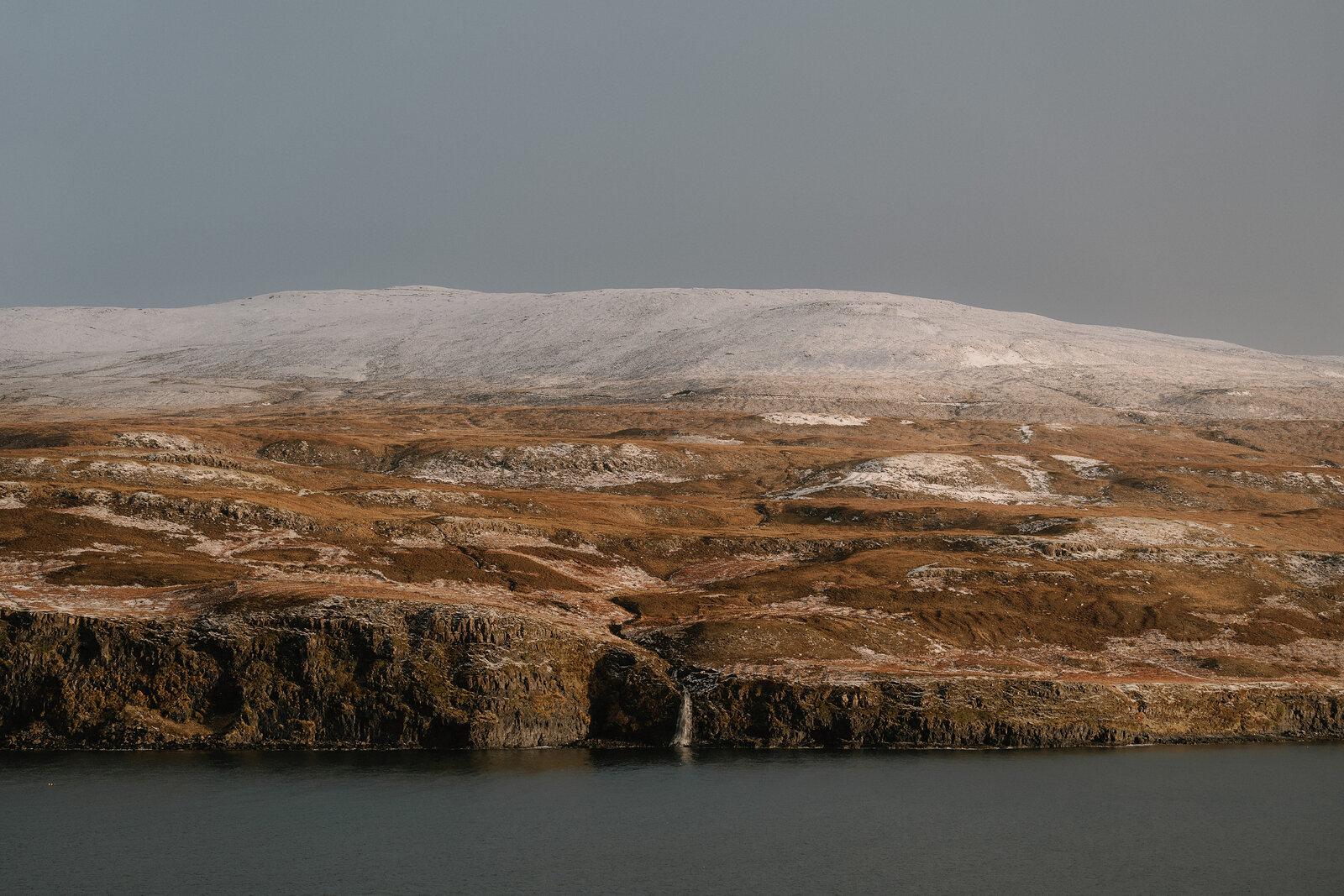 A waterfall drops down a low sea cliff below snow-dusted moorland on the Isle of Skye.