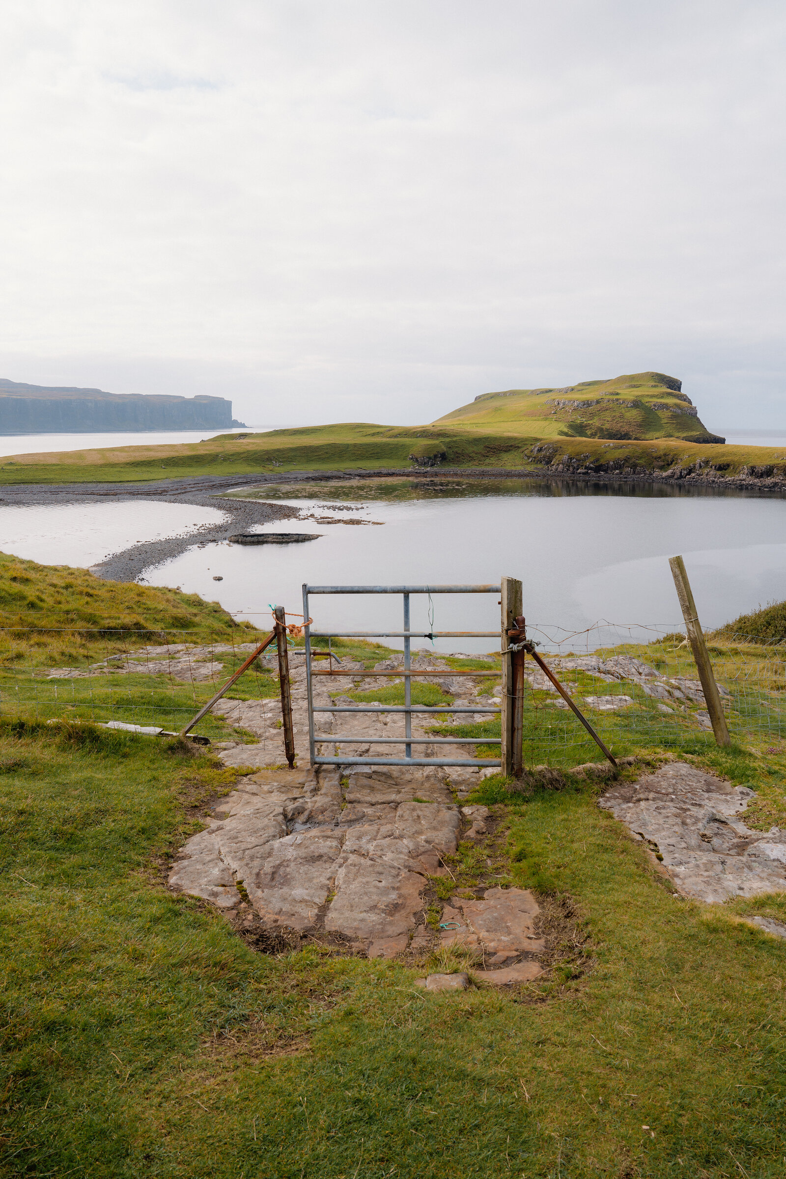 A metal gate opens onto a grassy tombolo headland enclosing a calm bay on the Isle of Skye.
