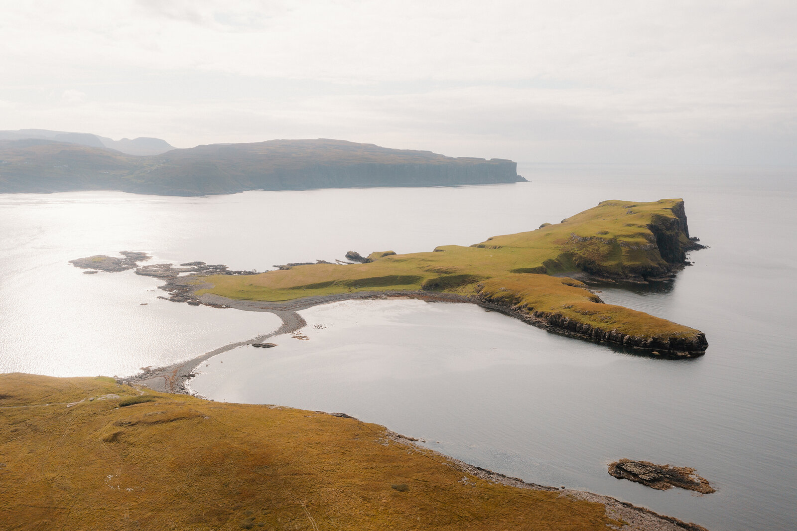 A tombolo headland reaches into calm water, seen from above on the Isle of Skye.