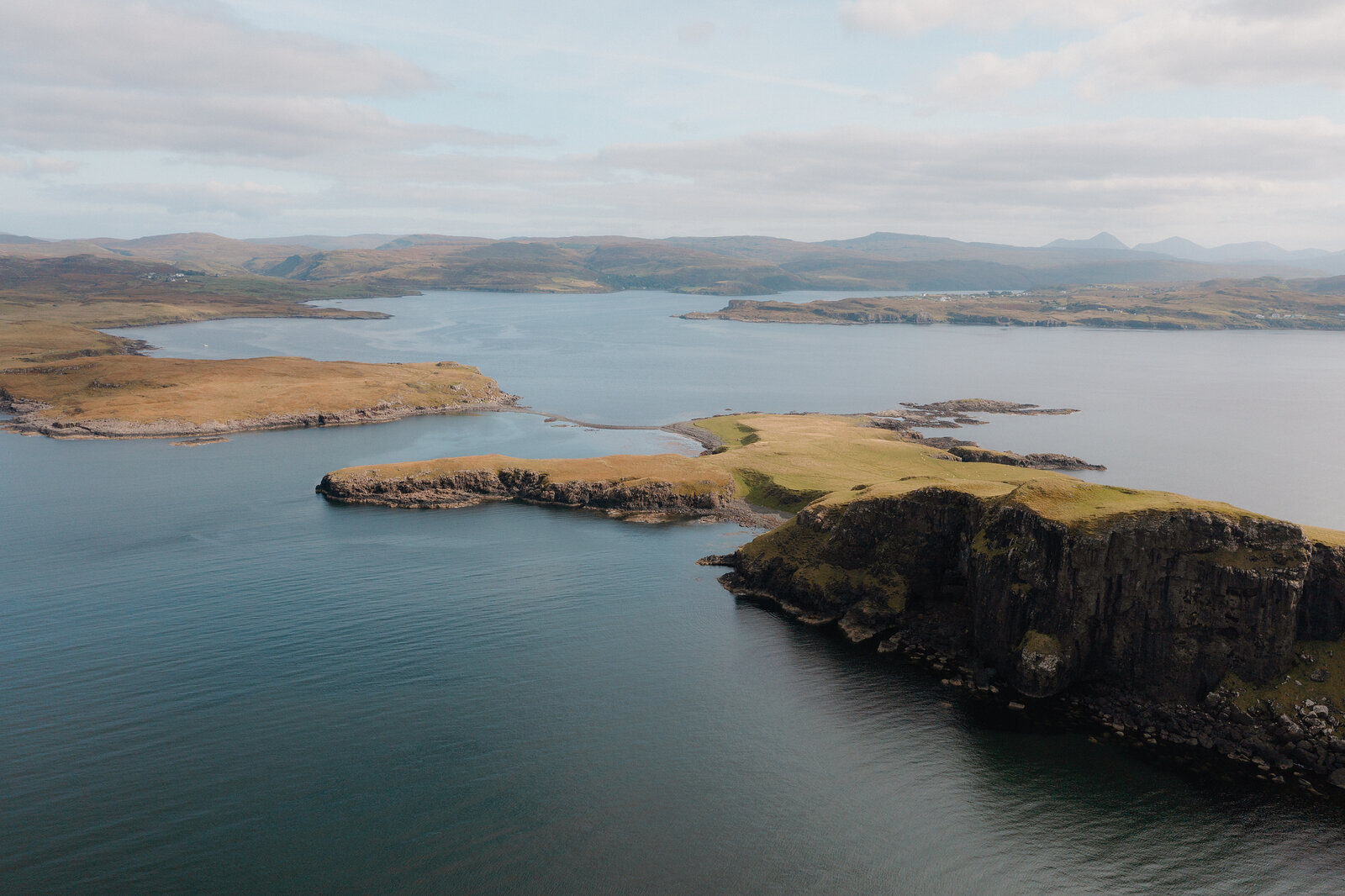 A grassy headland stretches into the sea with hills rising beyond, photographed from above on the Isle of Skye.