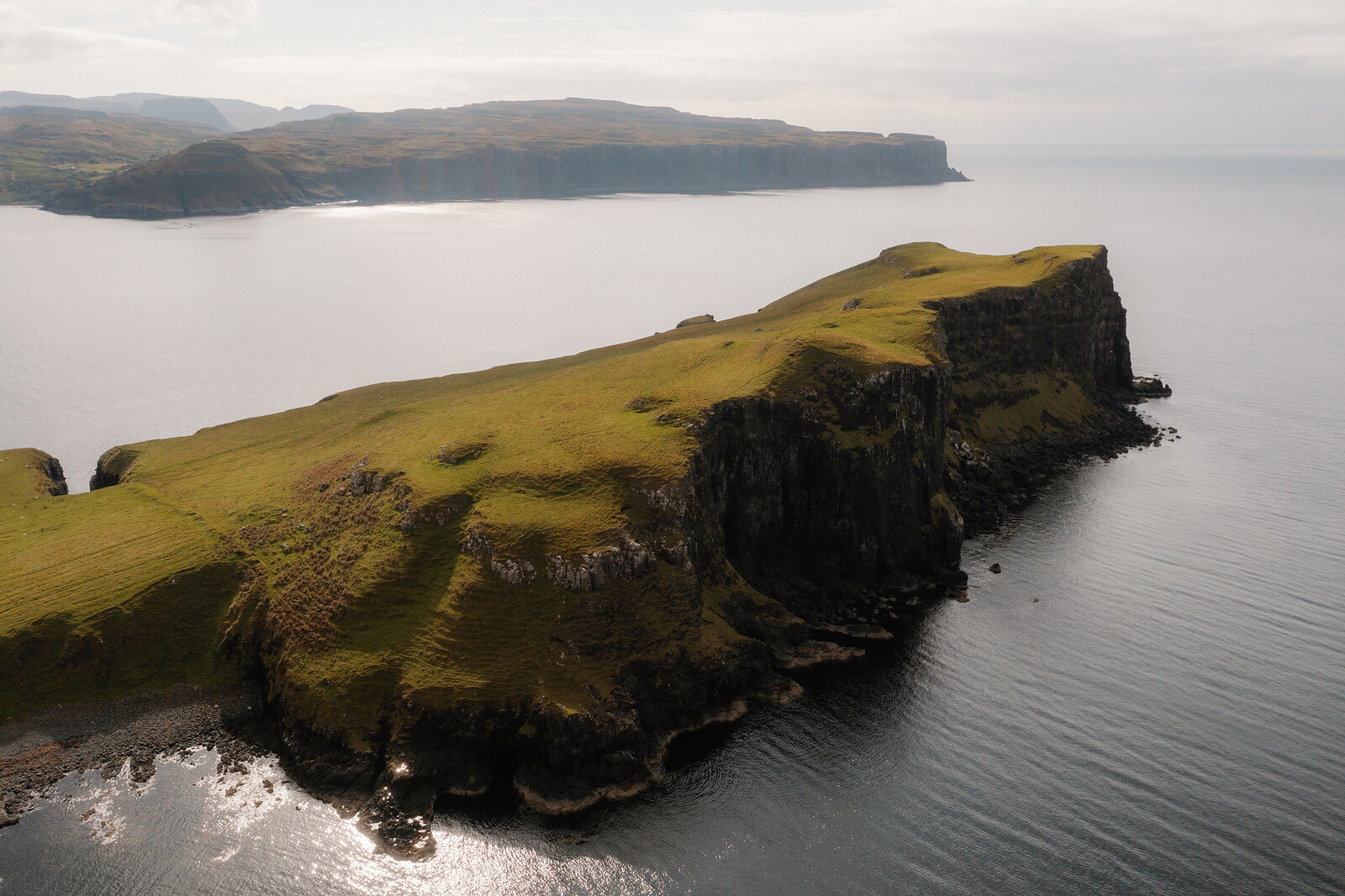 Dark cliffs edge a grassy headland reaching into a still sea, photographed from above on the Isle of Skye.