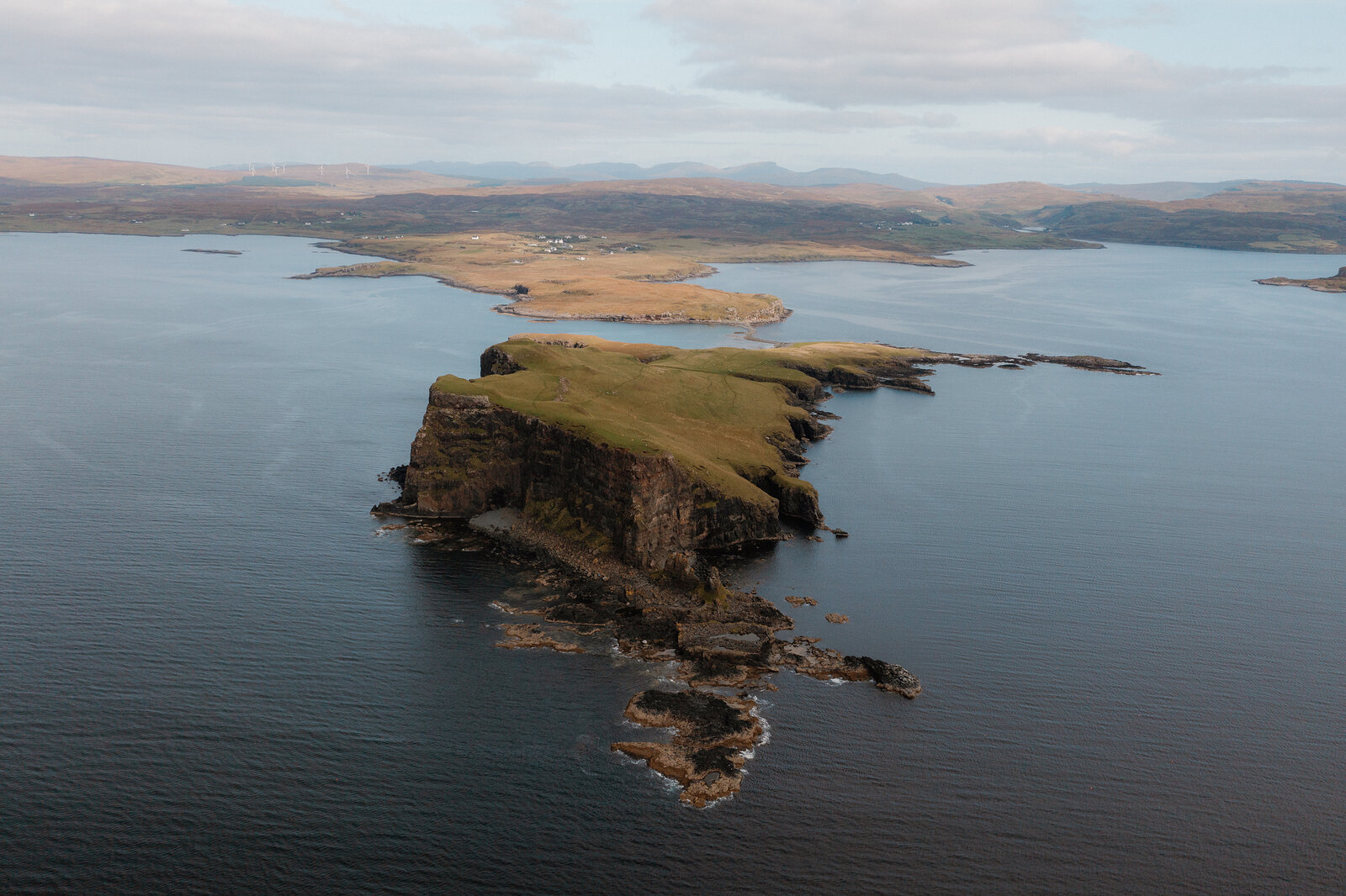 A tombolo headland with a small pool at its centre reaches into the sea, a village visible beyond on the Isle of Skye.