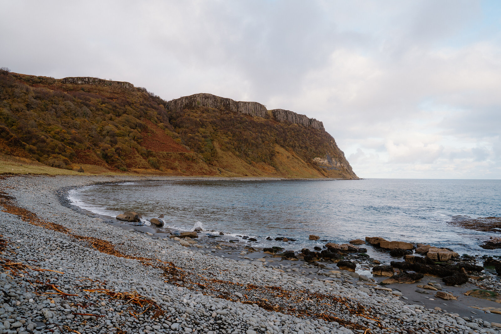 A pebble beach curving beneath autumn-coloured cliffs, the sea calm along the shore