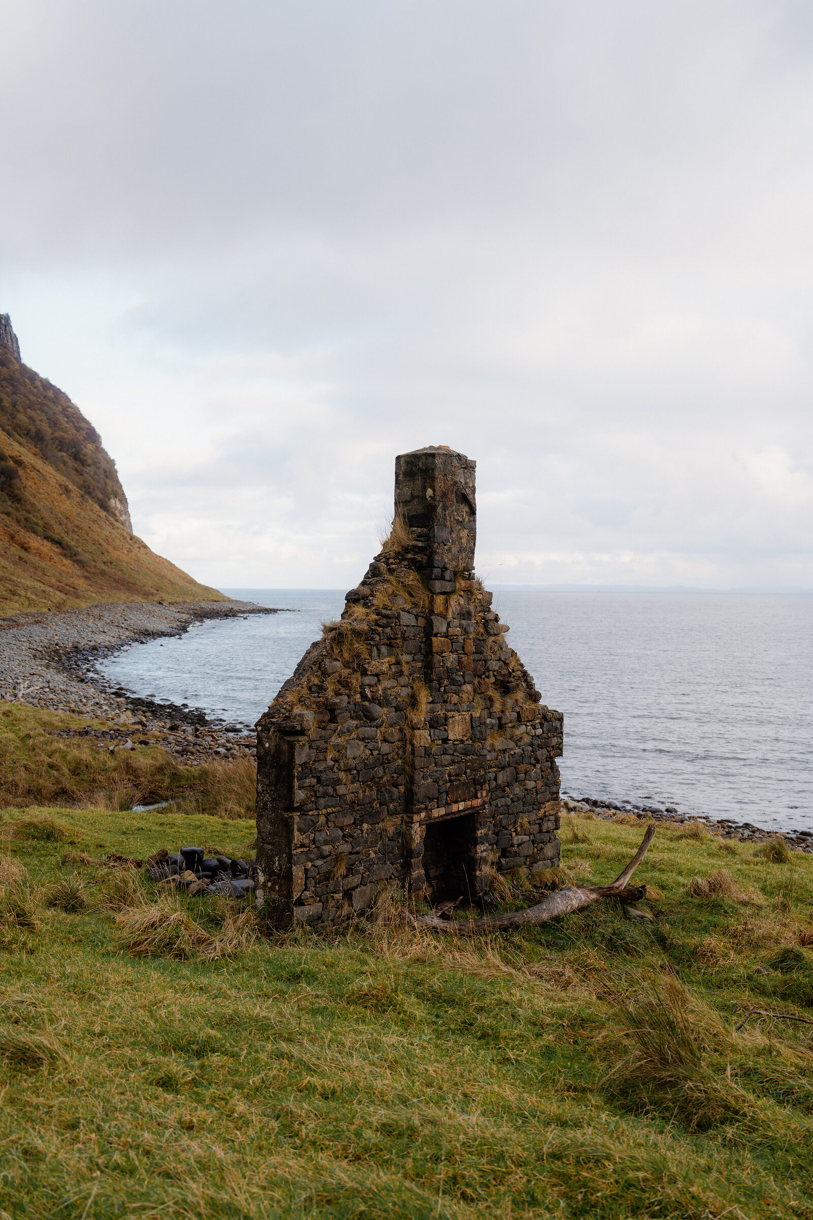 A roofless stone bothy with a tall chimney standing on a shoreline beneath steep hills