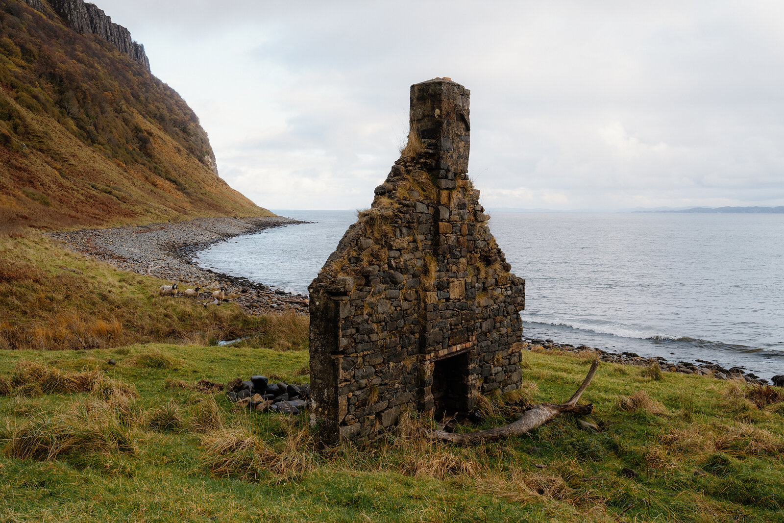 A roofless stone bothy with a tall chimney standing above a shingle shore beneath steep hills