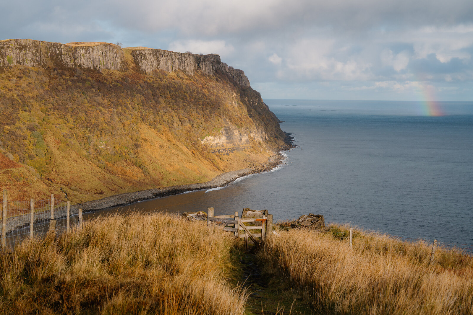 A faint rainbow arcing over the sea beside steep cliffs and a shingle bay, a gate set in the grass foreground