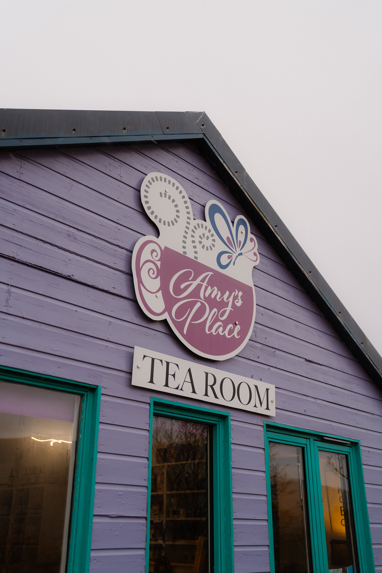 A sign shaped like a teacup with the words 'Amy's Place Tea Room' is mounted on the purple gable of a timber building.