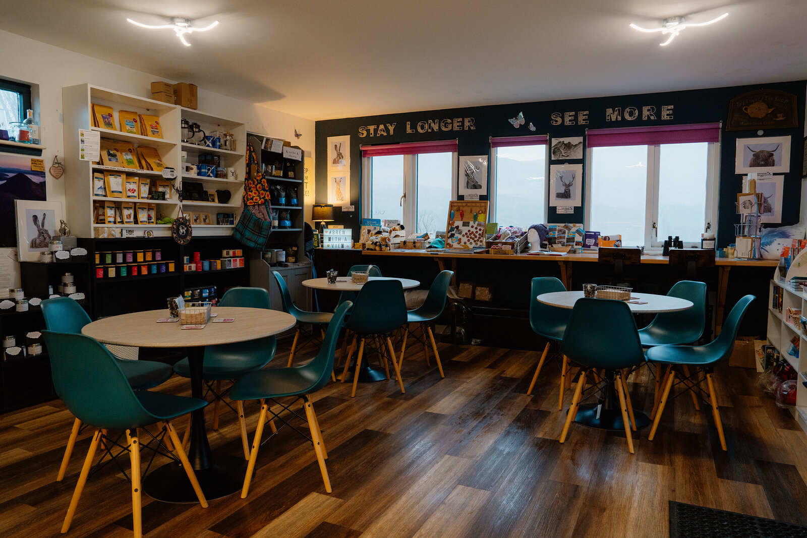 Round tables with teal chairs fill a tea room lined with gift shelves beneath a wall painted with 'Stay Longer, See More'.