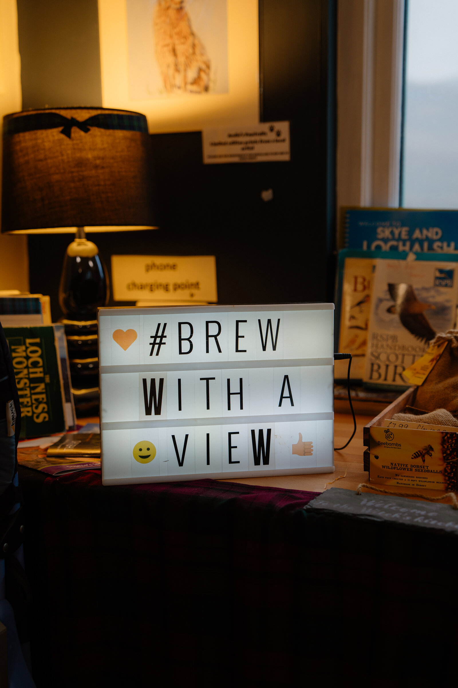A lightbox sign reading 'Brew with a view' sits on a counter beside a lamp, books and jars of local produce.