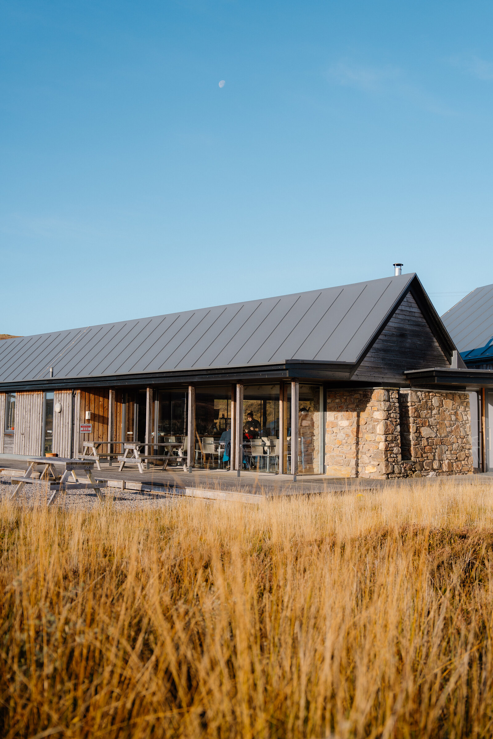 A timber and stone building with a pitched roof, diners visible inside beside picnic tables on the terrace