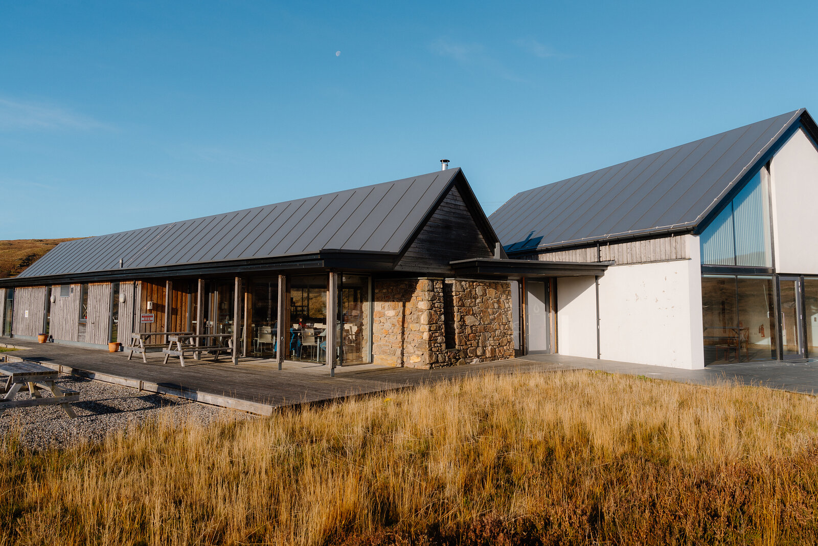 A modern timber and stone building with pitched roofs set among dune grass under a clear sky