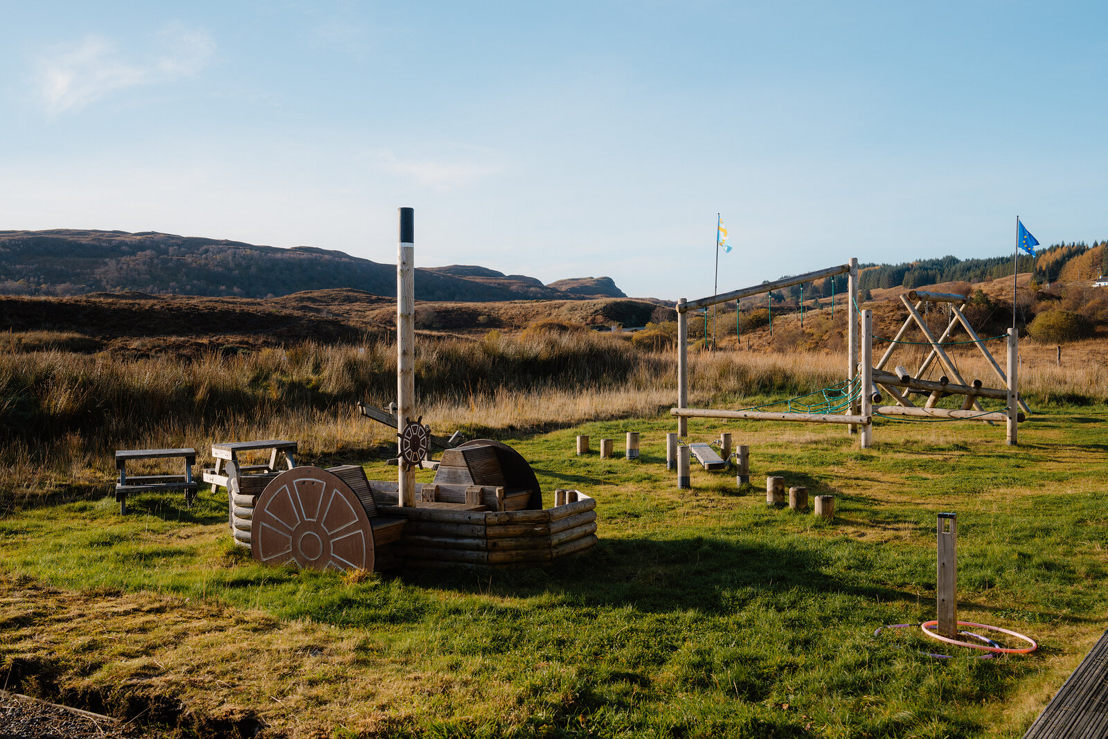 An outdoor play area with wooden climbing frames and flags set on grass, hills rising behind