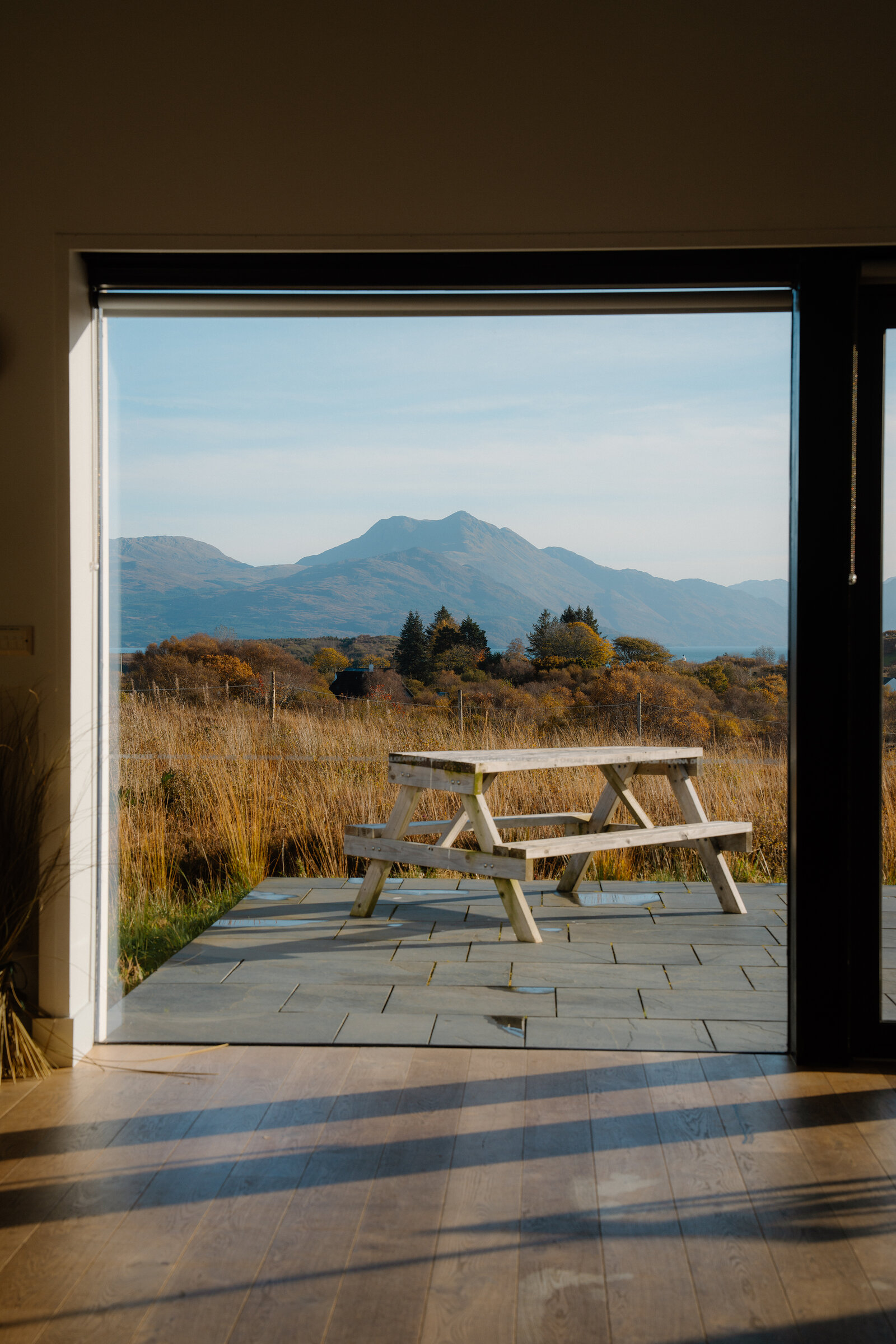 A wooden picnic table set on a stone patio beyond an open glass door, mountains rising in the distance