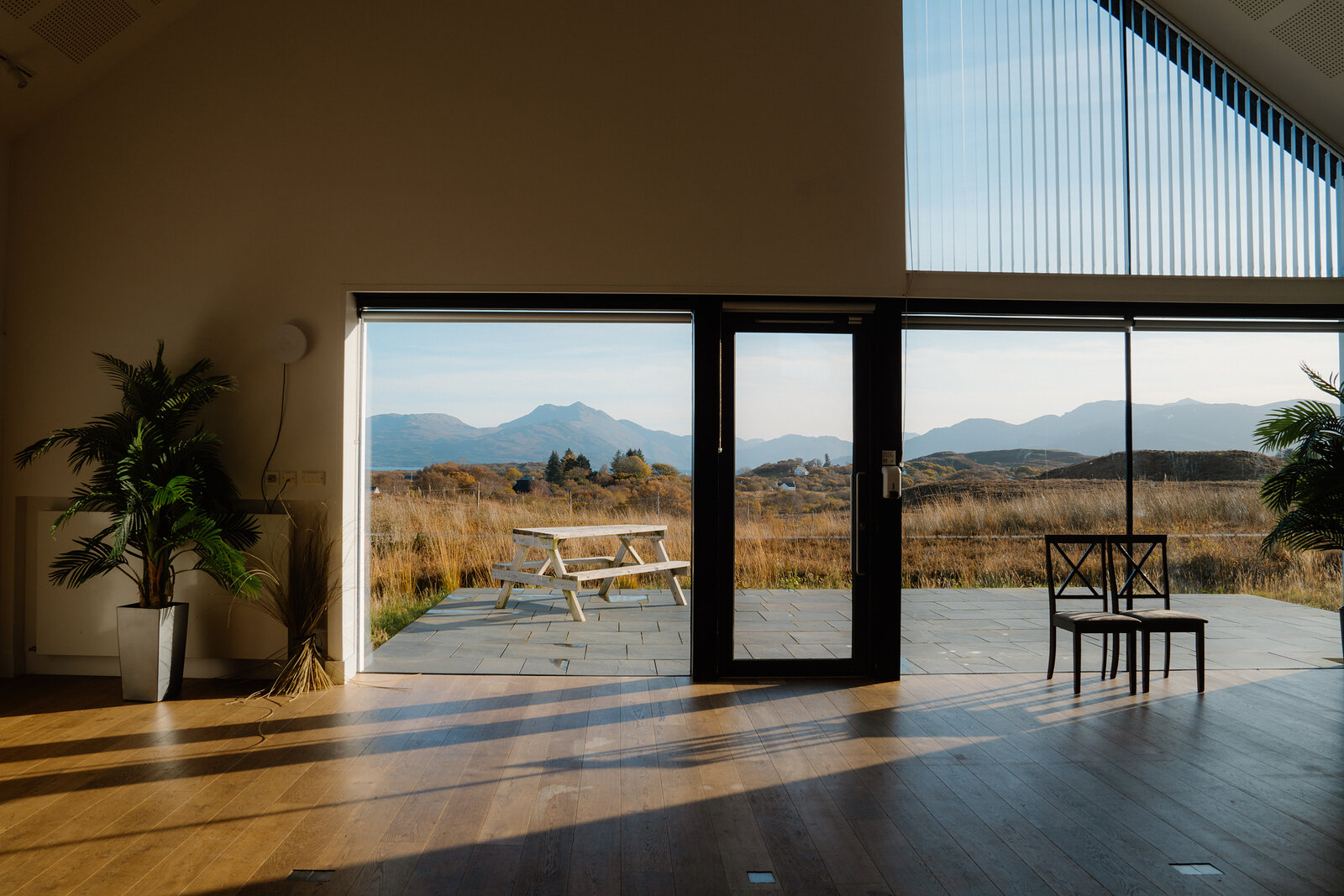A picnic table on a stone patio seen through open glass doors, mountains and moorland stretching beyond, houseplants and a wooden floor inside