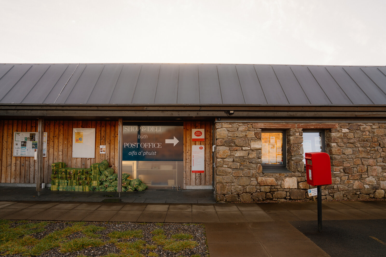 A stone and timber building with a sign reading Post Office, Oifis a' Phuist, beside a red postbox