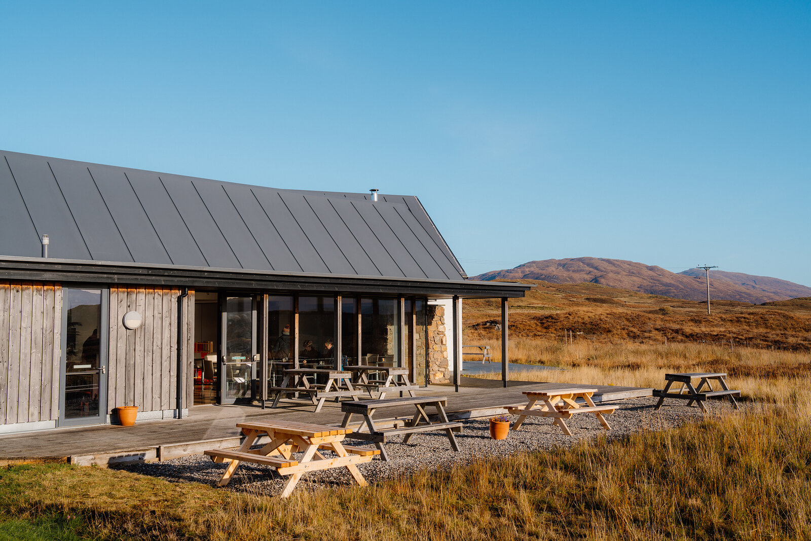 A timber-clad building with picnic tables set on gravel, hills rising behind under a clear sky