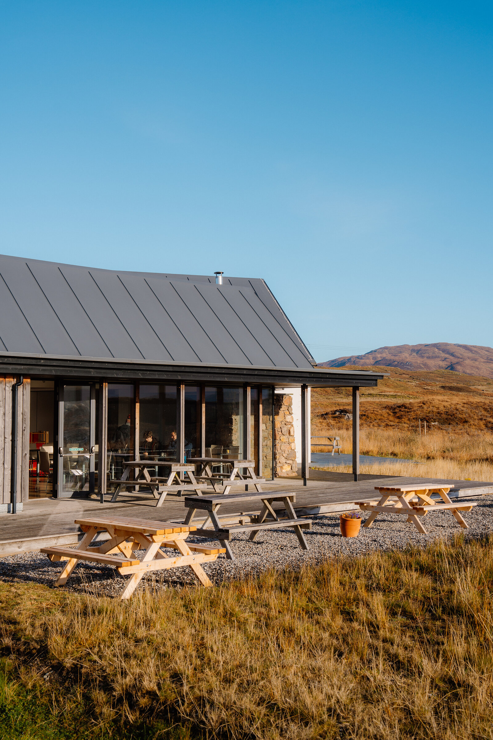 A dark-roofed timber building with picnic tables on its gravel terrace, hills rising behind under a clear sky