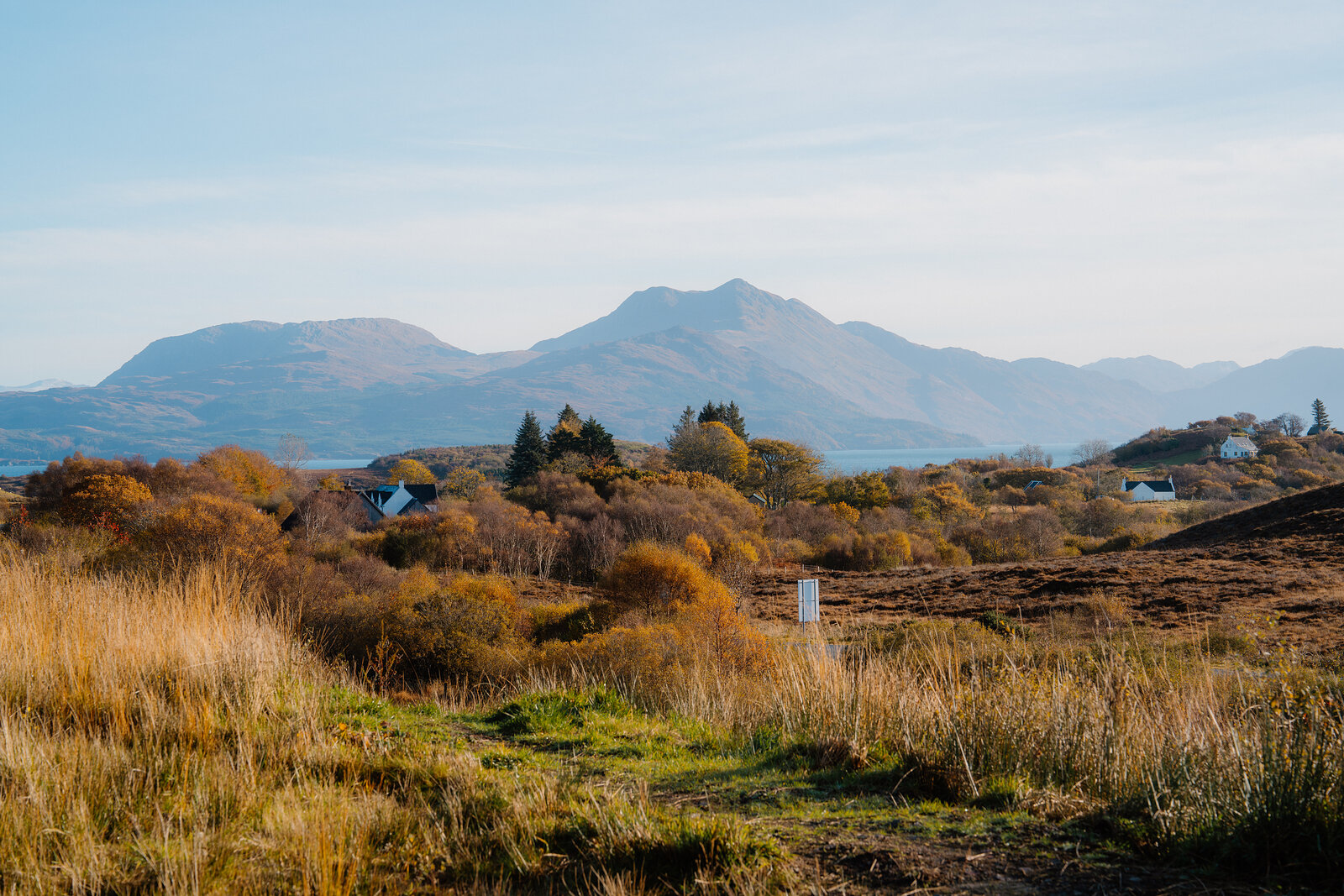 Cottages and autumn woodland set before mountains and a loch, a marker post standing in the foreground