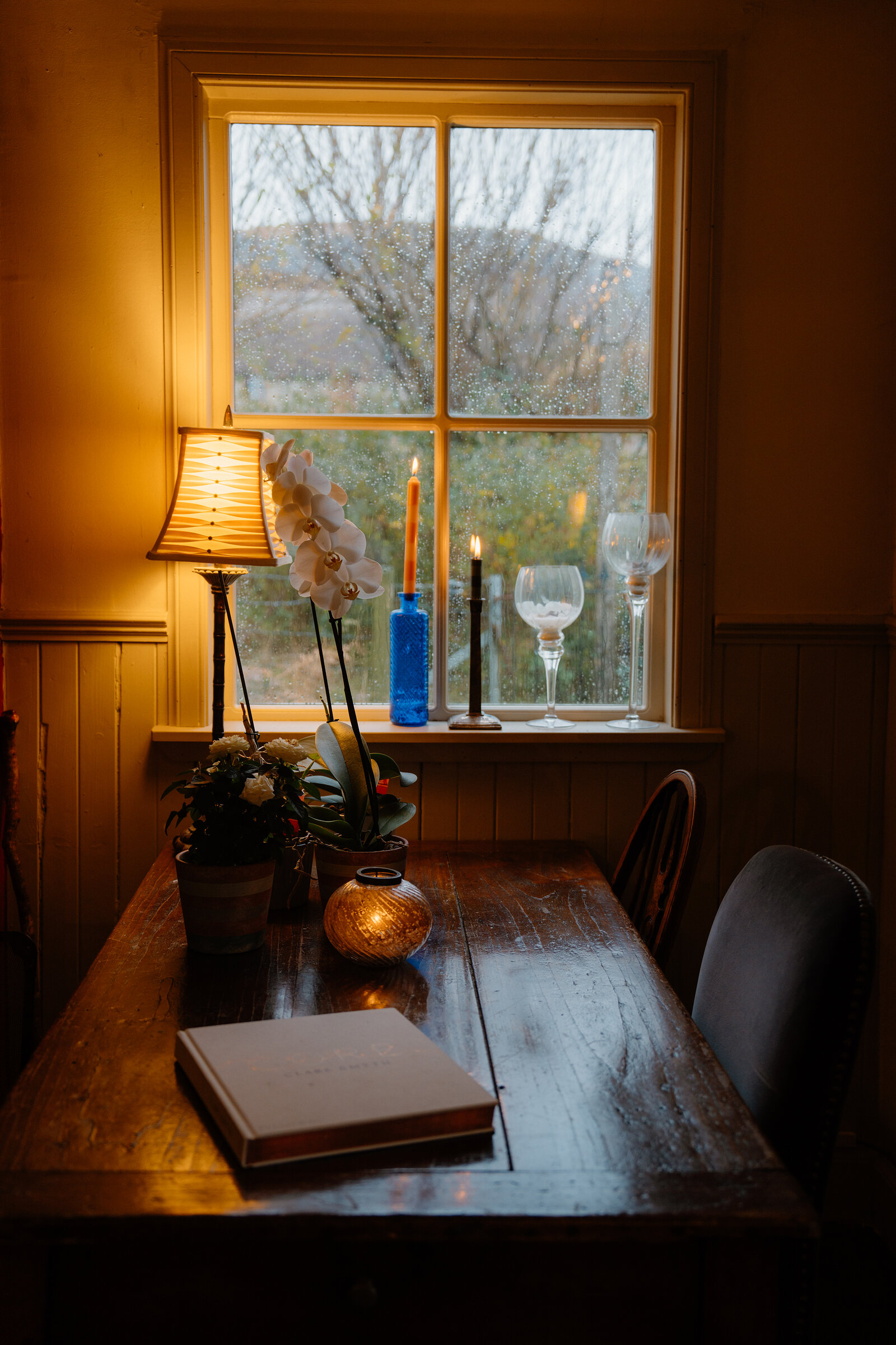 A wooden table at Coruisk House holds a lit candle, an orchid and wine glasses beside a rain-streaked window.