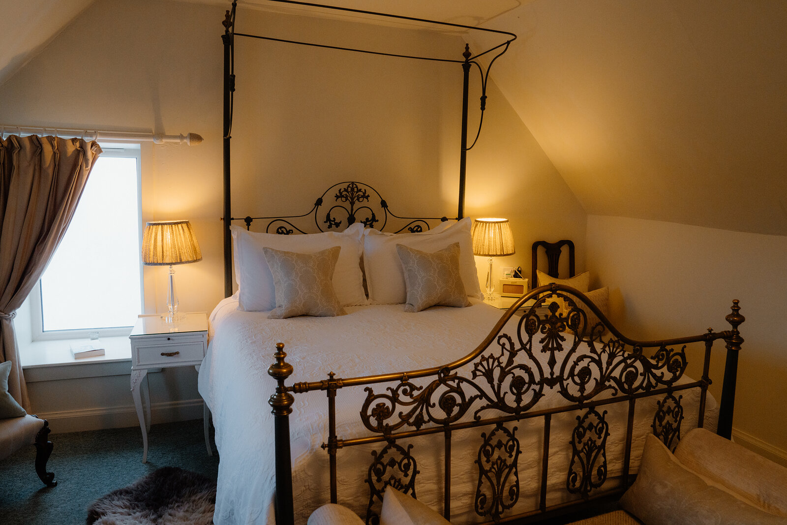 A wrought-iron four-poster bed dressed in white linen and cushions stands beneath a sloped ceiling at Coruisk House.