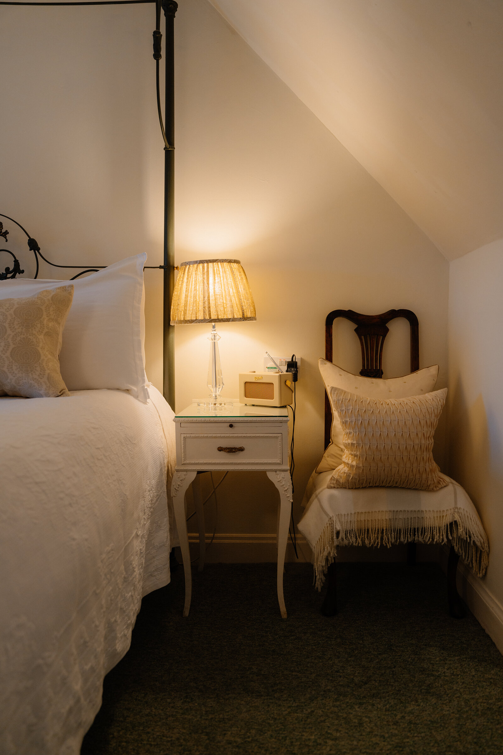 A lamp-lit bedside table and an upholstered chair with cushions sit beside a wrought-iron bed at Coruisk House.