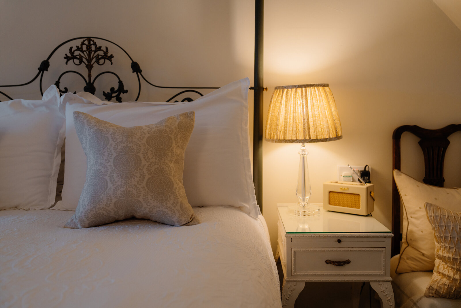 A lamp lights a bedside table beside a wrought-iron bed dressed with patterned cushions at Coruisk House.