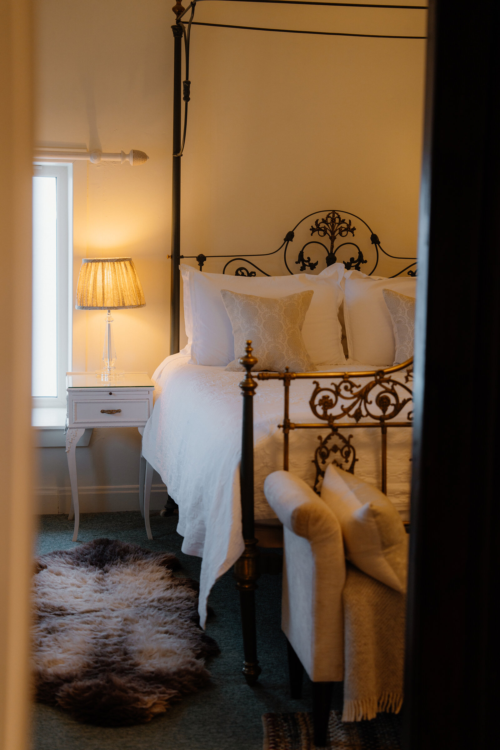 A wrought-iron four-poster bed with cream cushions stands beside an upholstered armchair and a sheepskin rug at Coruisk House.