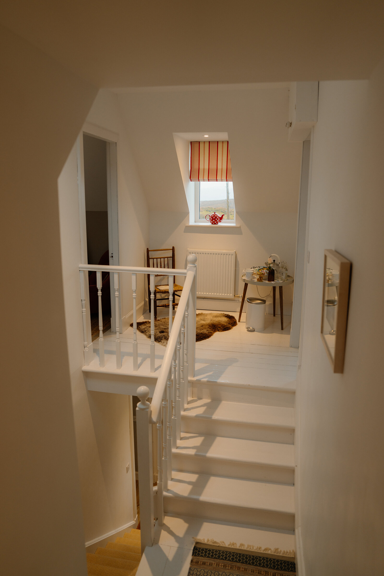 A white-painted staircase descends toward a landing furnished with a chair, a sheepskin rug and a striped roller blind at Coruisk House.