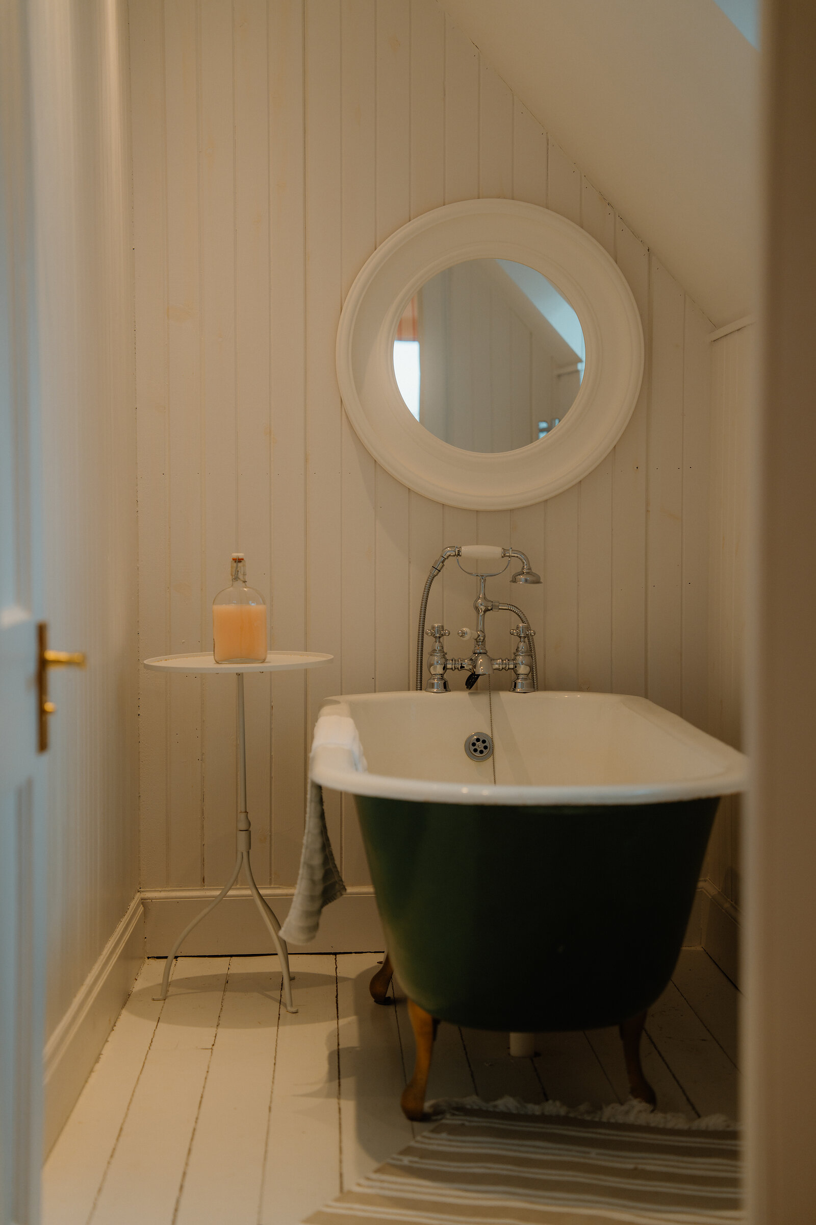 A green clawfoot bathtub stands below a round mirror beside a side table holding a bottle of bath oil at Coruisk House.