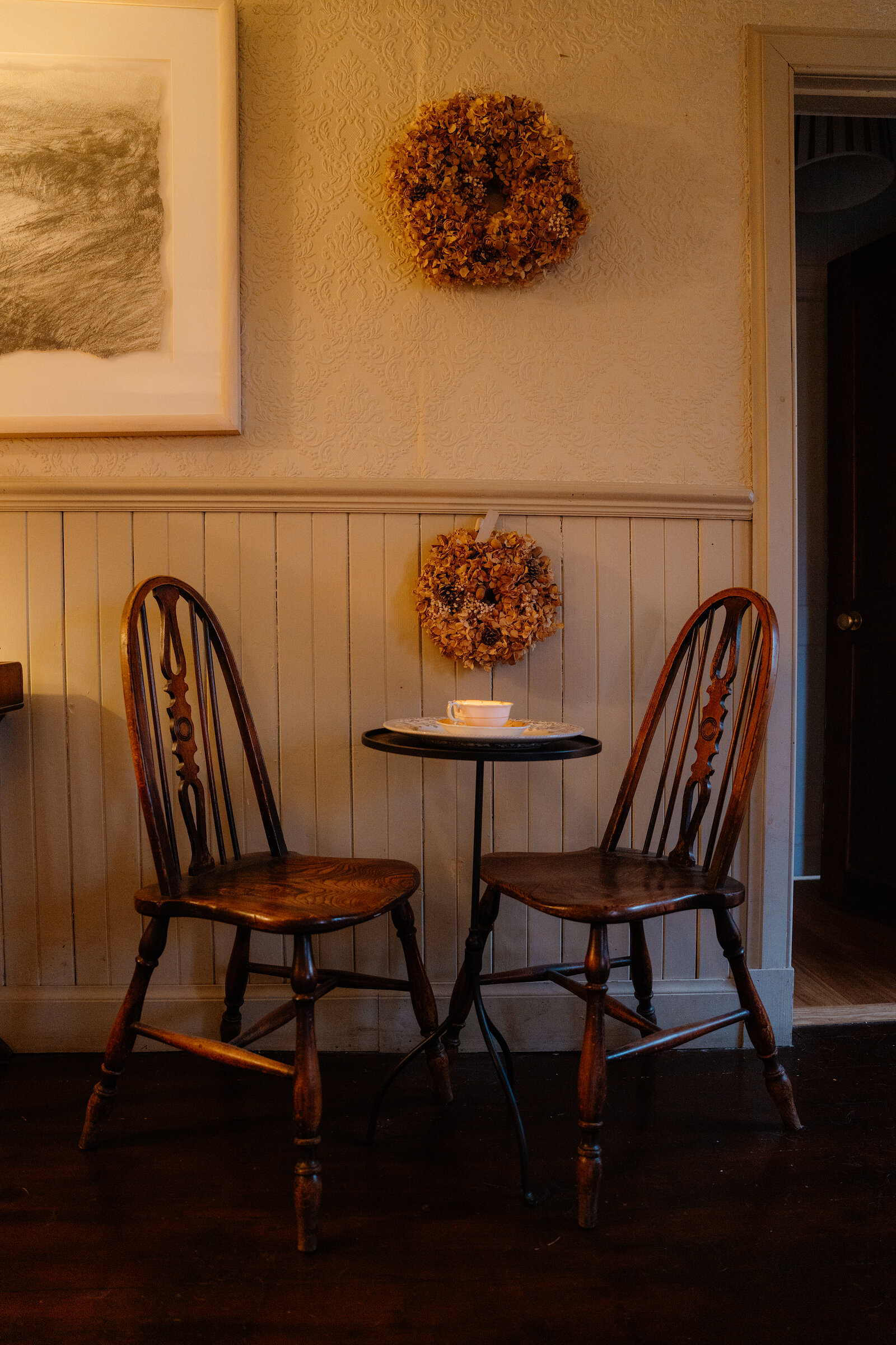 Two wooden chairs flank a small round table set with a cup and saucer beneath dried flower wreaths on the wall at Coruisk House.