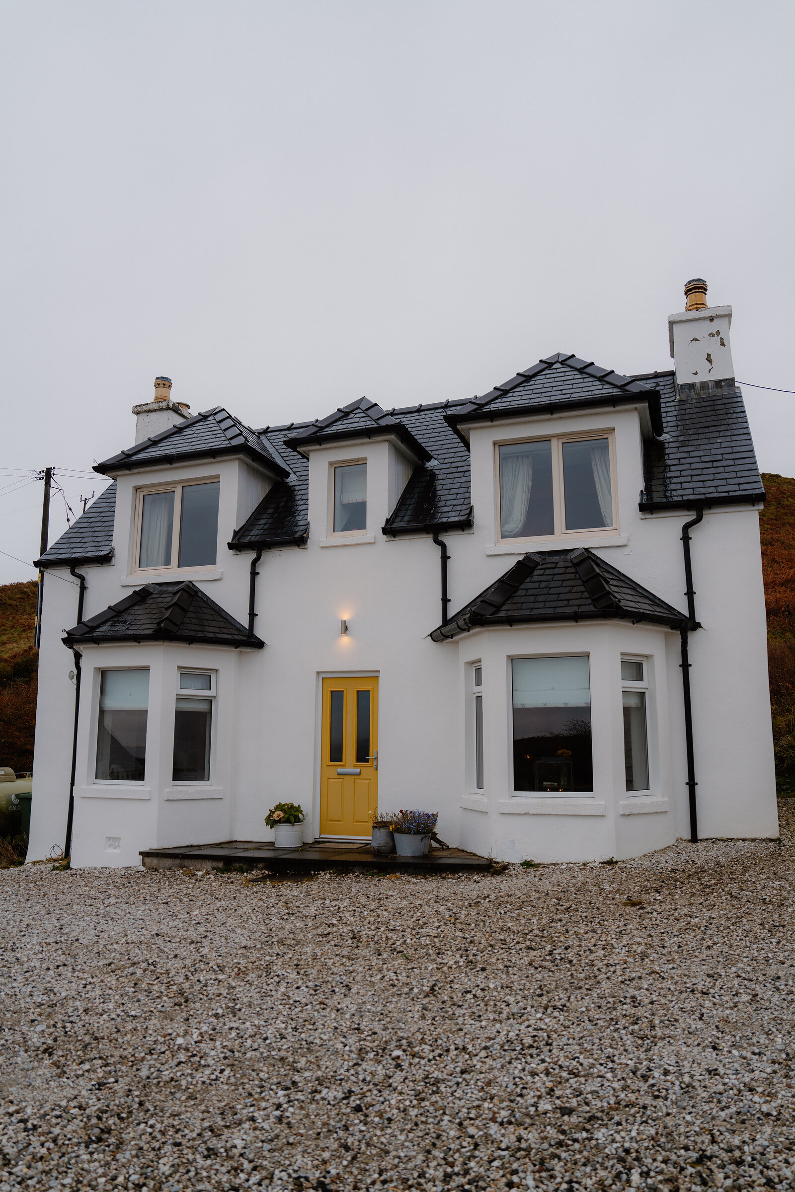 Coruisk House's white exterior features a yellow front door, three gabled dormers and chimneys against a grey sky.