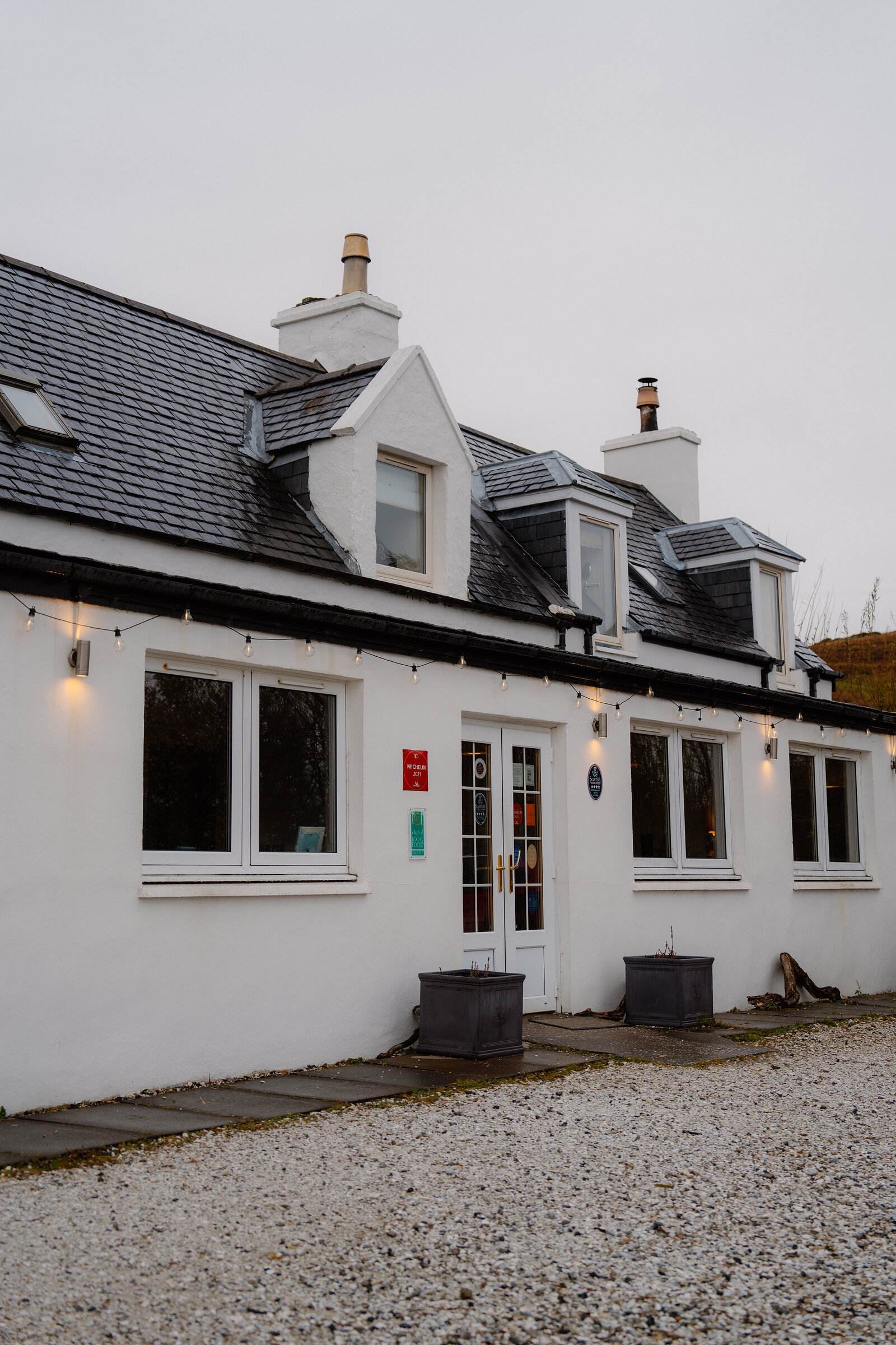 Coruisk House's low white building is strung with outdoor lights along its eaves above a gravel path.