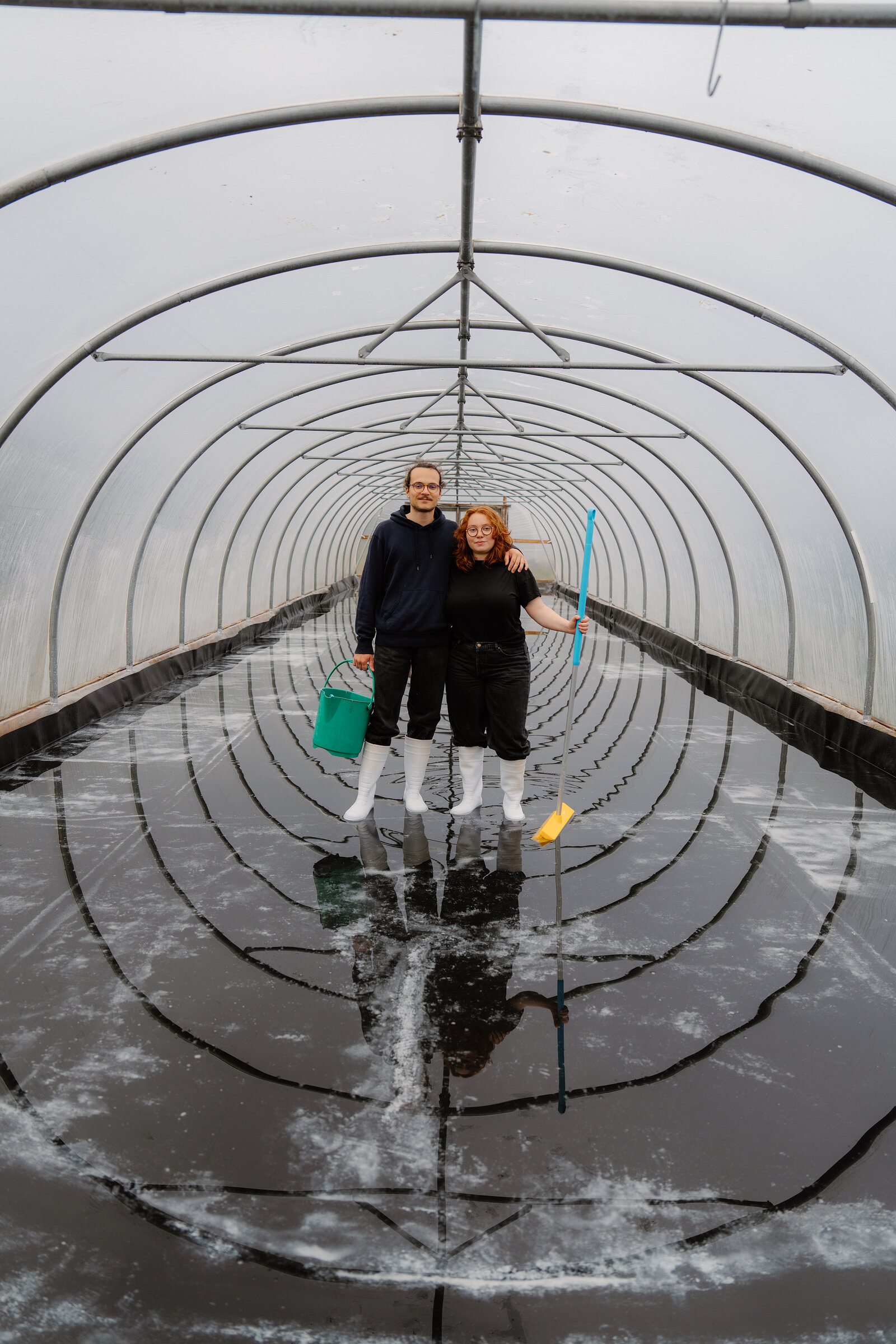 A couple in white boots stand together holding a bucket and squeegee inside a seawater polytunnel at Isle of Skye Sea Salt Company.