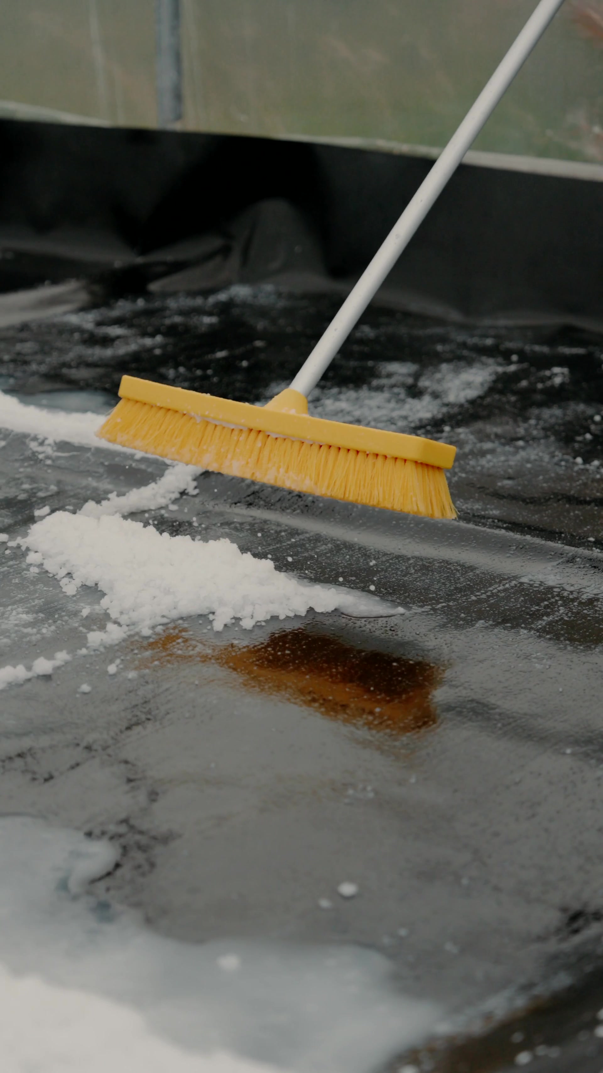 A yellow-bristled broom sweeps salt crystals across a black-lined polytunnel floor, a rust-coloured stain marking the surface beneath.