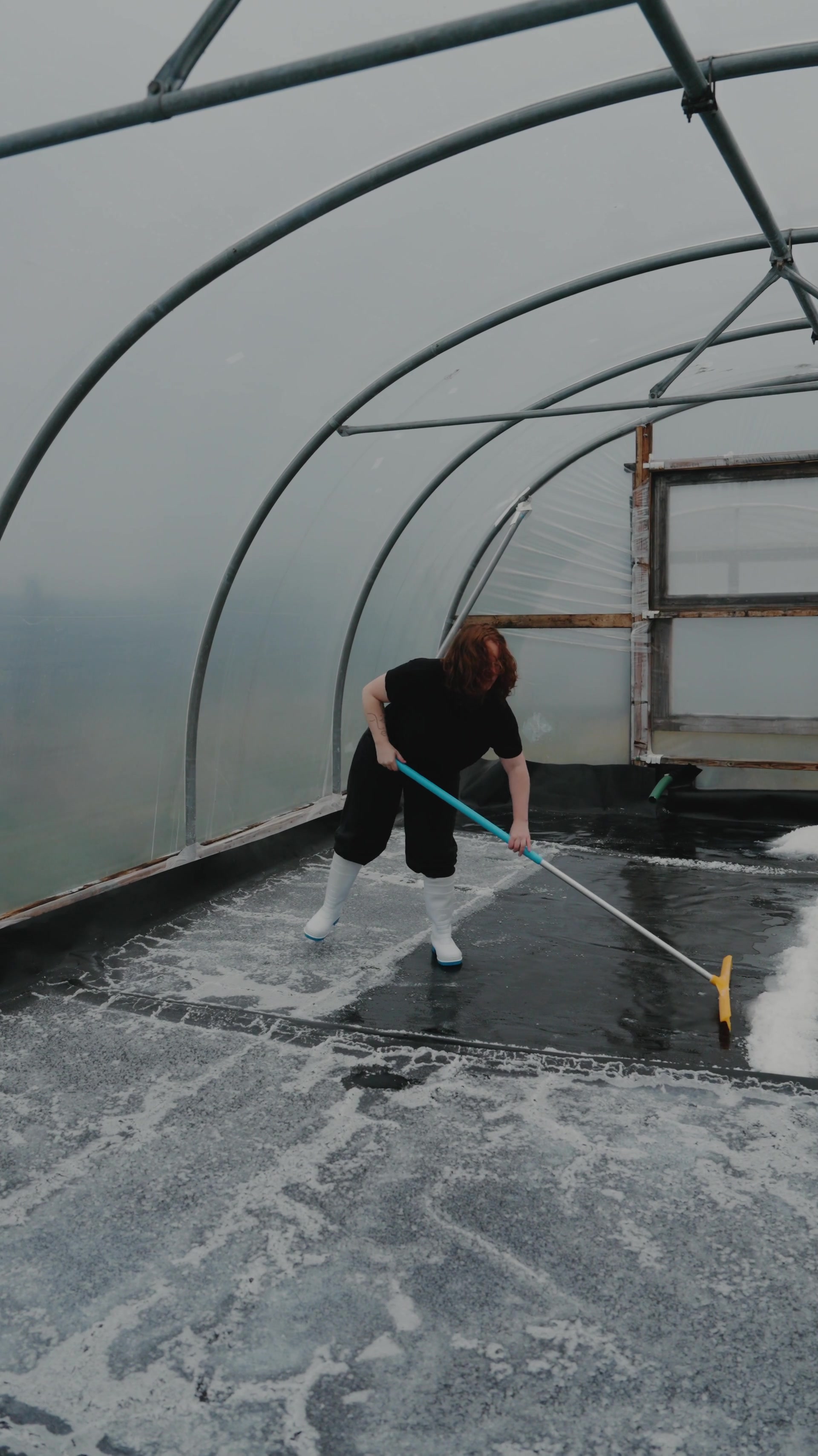 A woman in wellies sweeps a squeegee across a frosted polytunnel floor, ice and snow streaked across the black lining.
