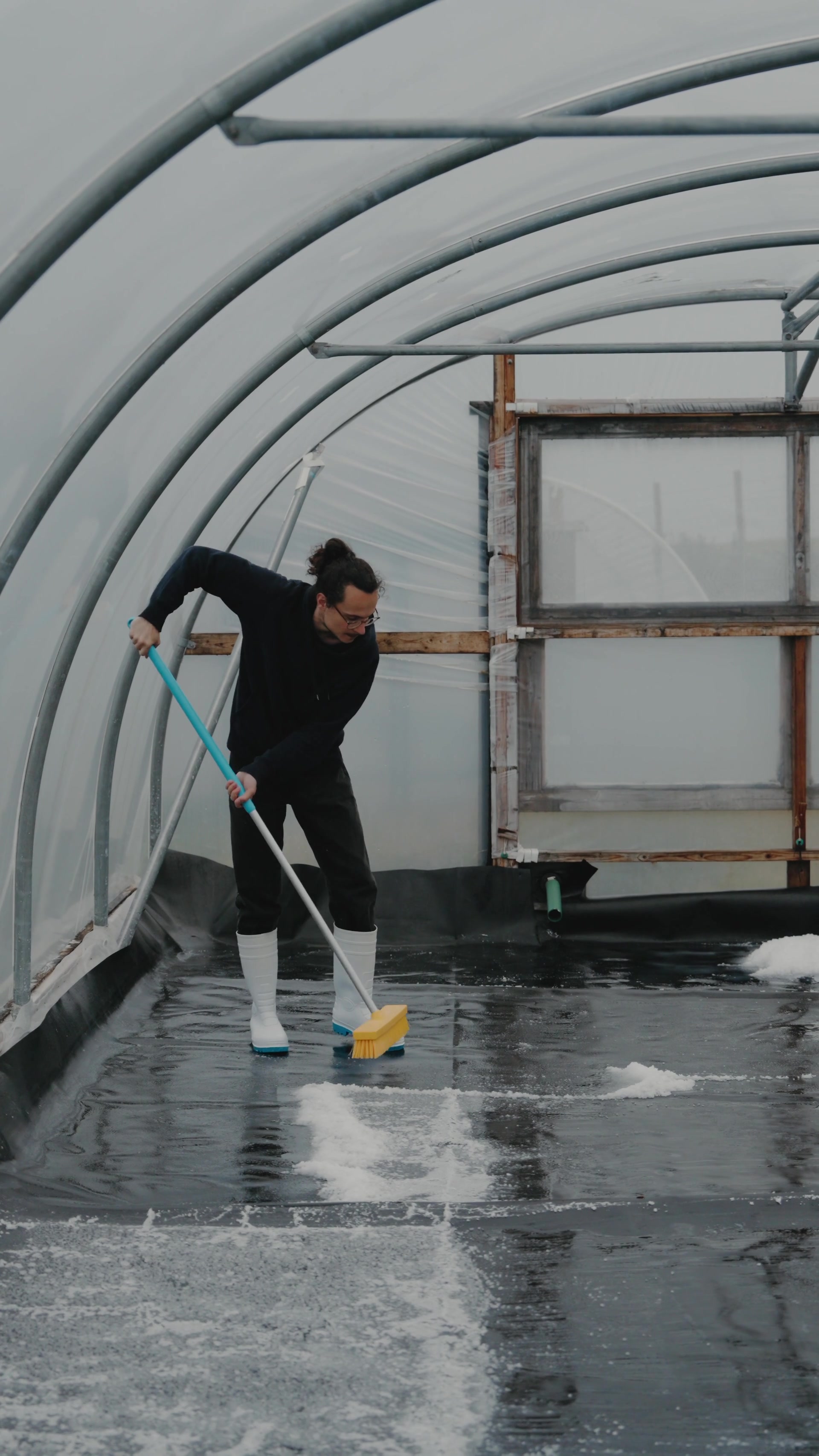 A man in wellies scrubs a frosted polytunnel floor with a squeegee broom, patches of snow streaked across the black lining.