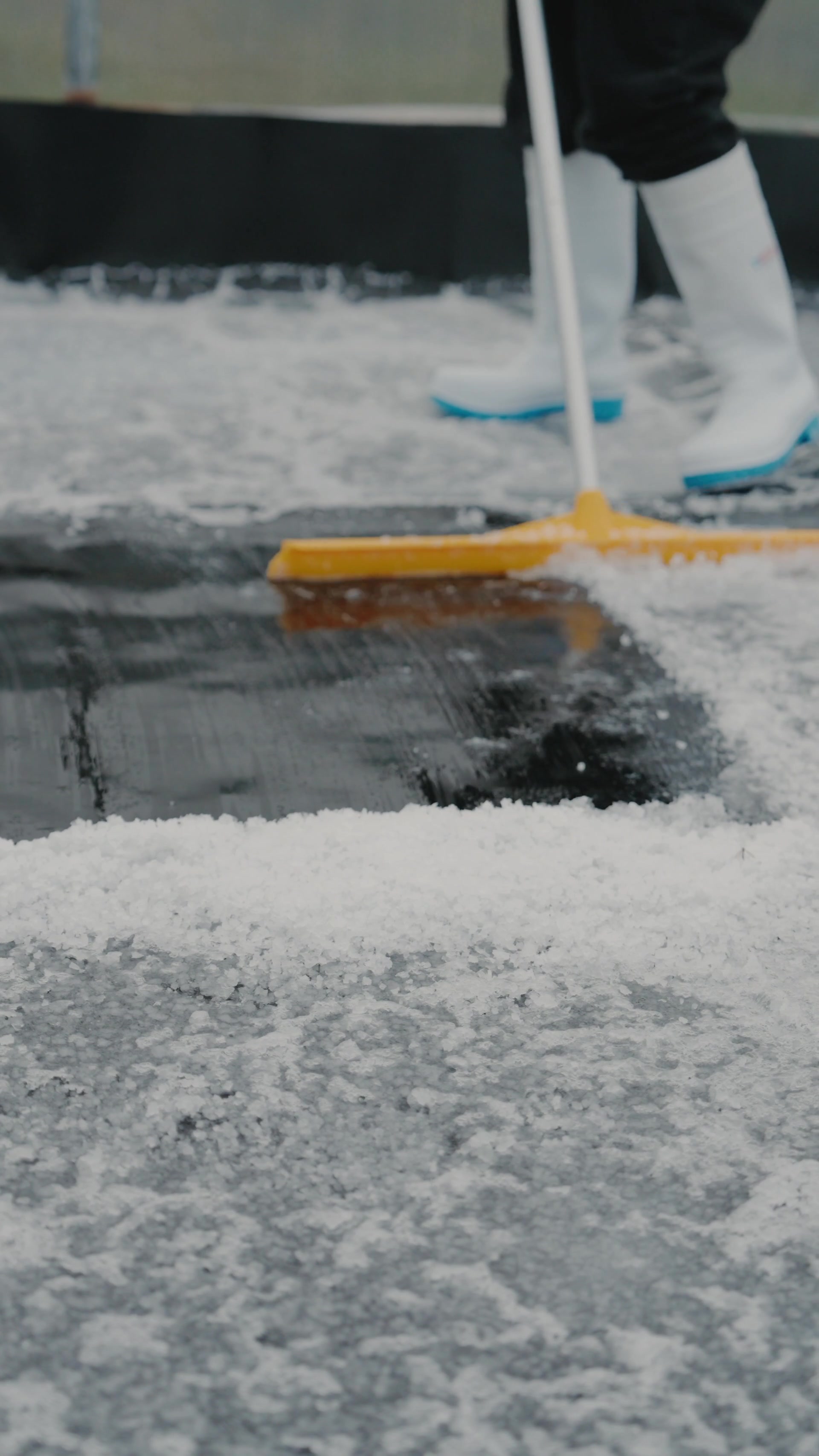 A person in white wellies pushes a yellow squeegee broom across a frosted polytunnel floor, ridges of salted ice piled at its edge.